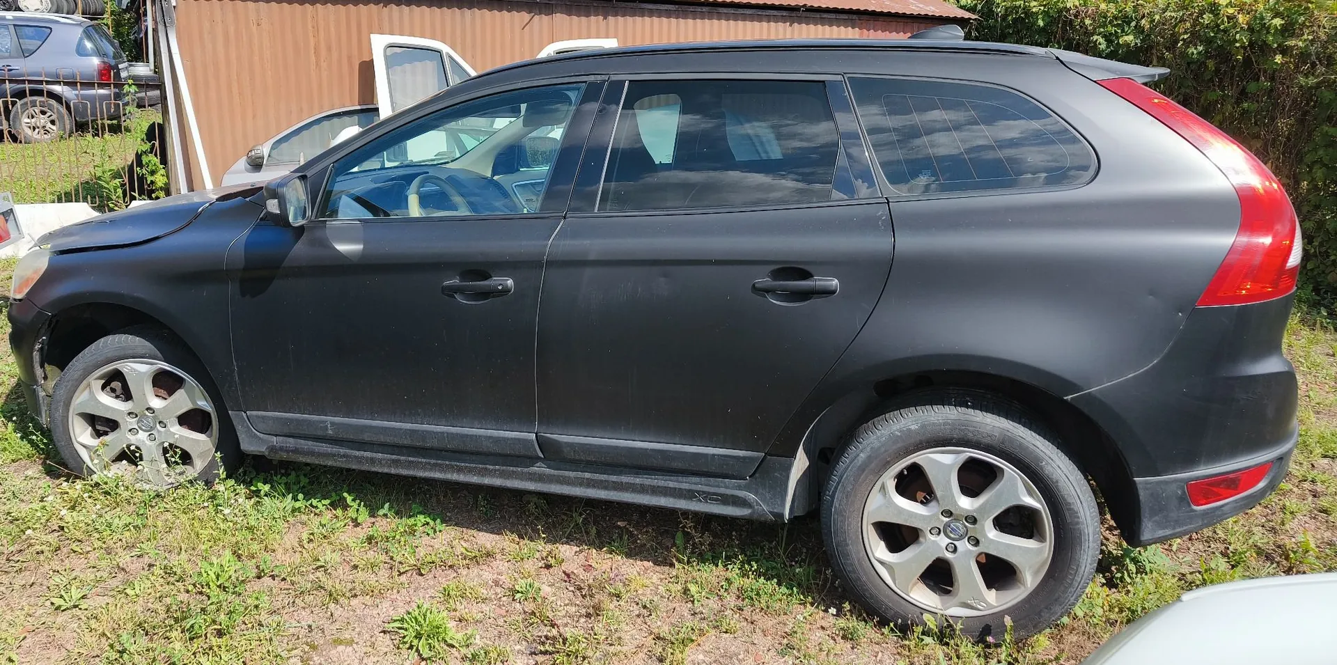 Czarny samochód typu kombi zaparkowany na trawiastym podłożu. Pojazd ma matowy lakier i jest widoczny z profilu od strony pasażera. W tle widać fragmenty innych samochodów oraz brązową ścianę budynku.