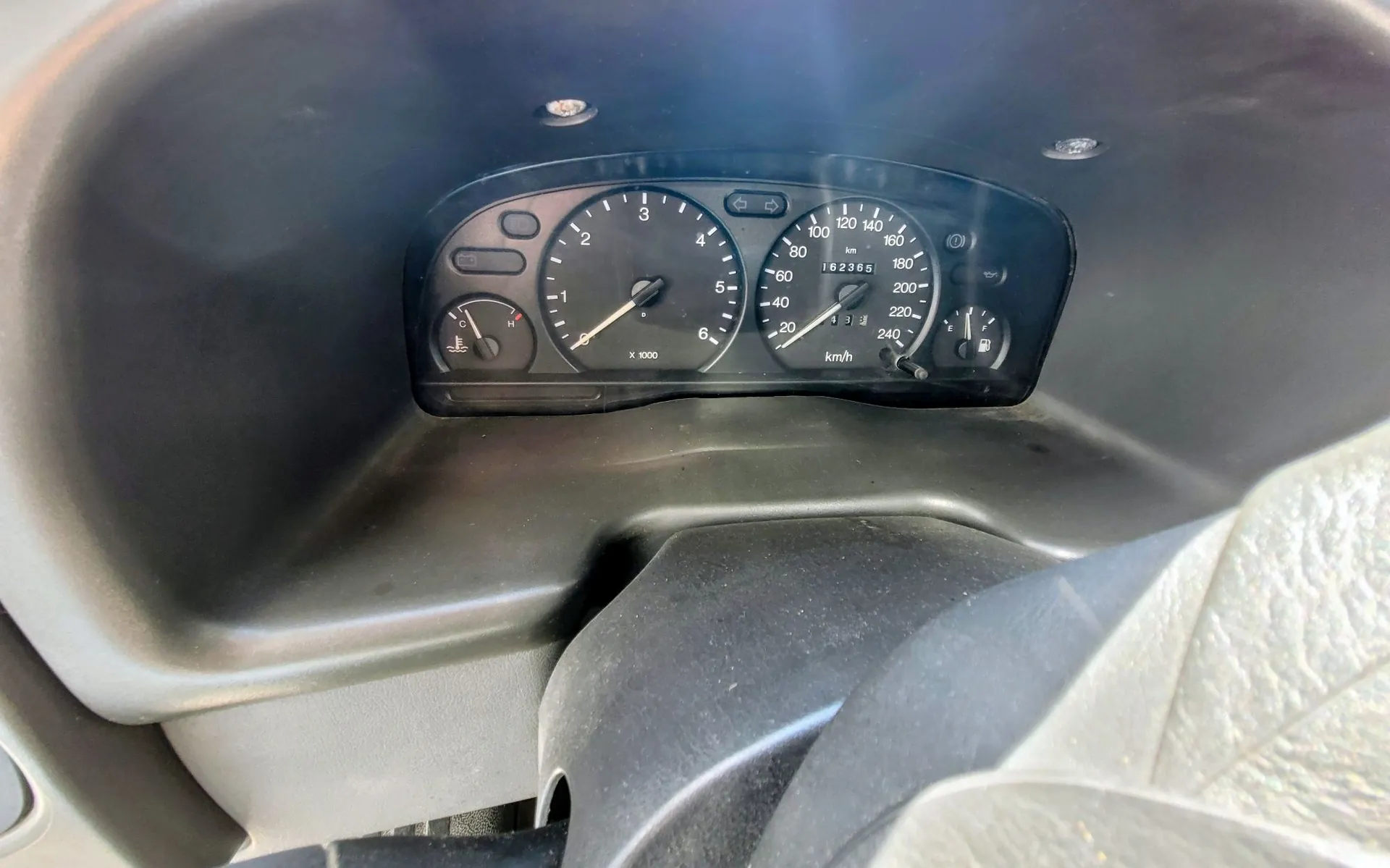 Zbliżenie na panel instrumentów samochodu. Widoczny jest prędkościomierz z maksymalną wartością 240 km/h, obrotomierz z oznaczeniem X 1000 oraz wskaźniki paliwa i temperatury. Licznik przebiegu wskazuje wartość 162365. Panel jest osadzony w ciemnoszarej obudowie deski rozdzielczej.