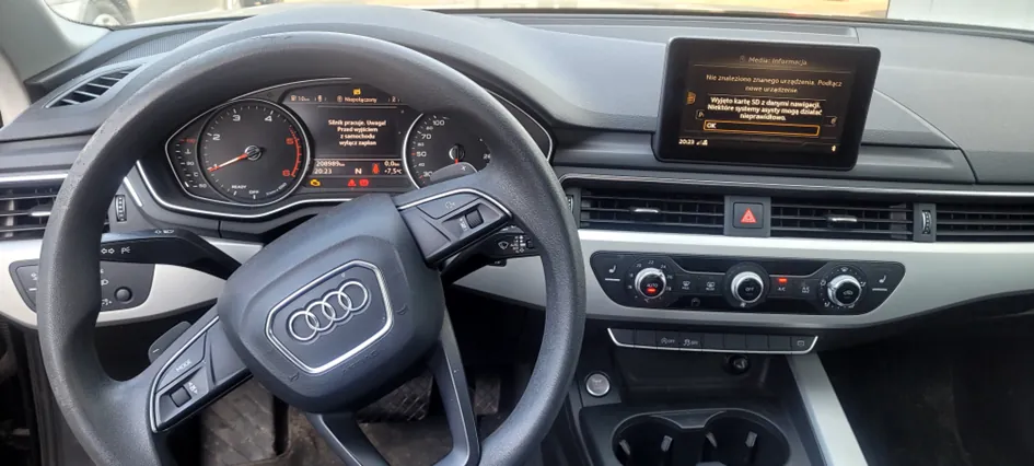 Wnętrze samochodu marki Audi z widoczną kierownicą z logo Audi, tablicą rozdzielczą z prędkościomierzem i tachometrem, centralnym ekranem wyświetlającym komunikat w języku polskim: "Nie ma żadnego zapisanego urządzenia. Podłącz nowe urządzenie. Wybierz kartę SD z danymi nawigacji. Niektóre systemy mogą nie działać prawidłowo." oraz panelami klimatyzacji i sterowania. Czas na ekranie: 20:21.