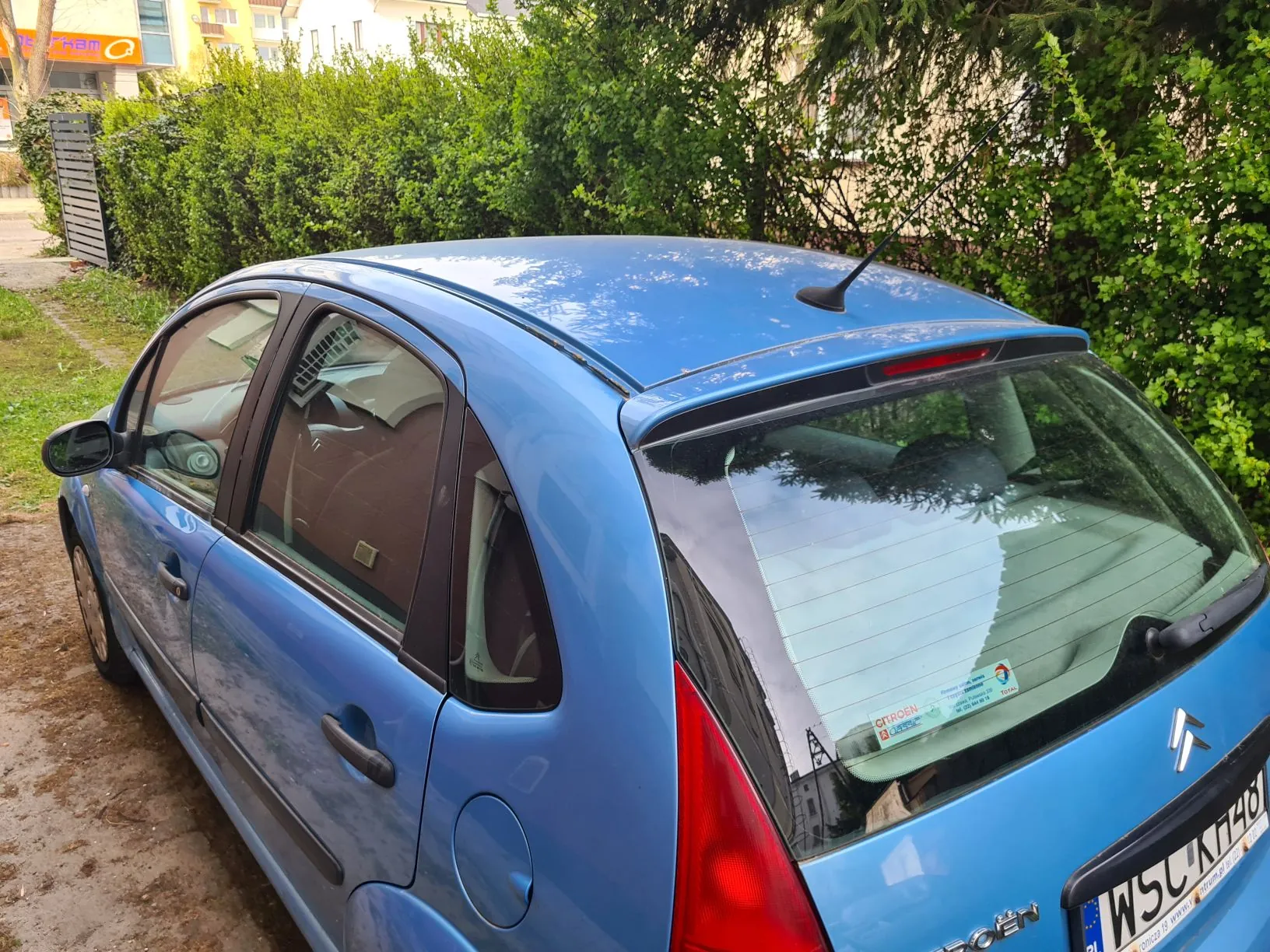 Niebieski samochód marki Citroën zaparkowany na brudnej drodze. Widok z tyłu i boku. Na szybie tylnej widoczne naklejki, w tym z napisem "CITROËN" oraz tablica rejestracyjna WSC KH48. W tle zielone krzewy, drzewa i budynki mieszkalne z częściowo widocznym pomarańczowym szyldem. Samochód ma czarną antenę i czerwone światła tylne.