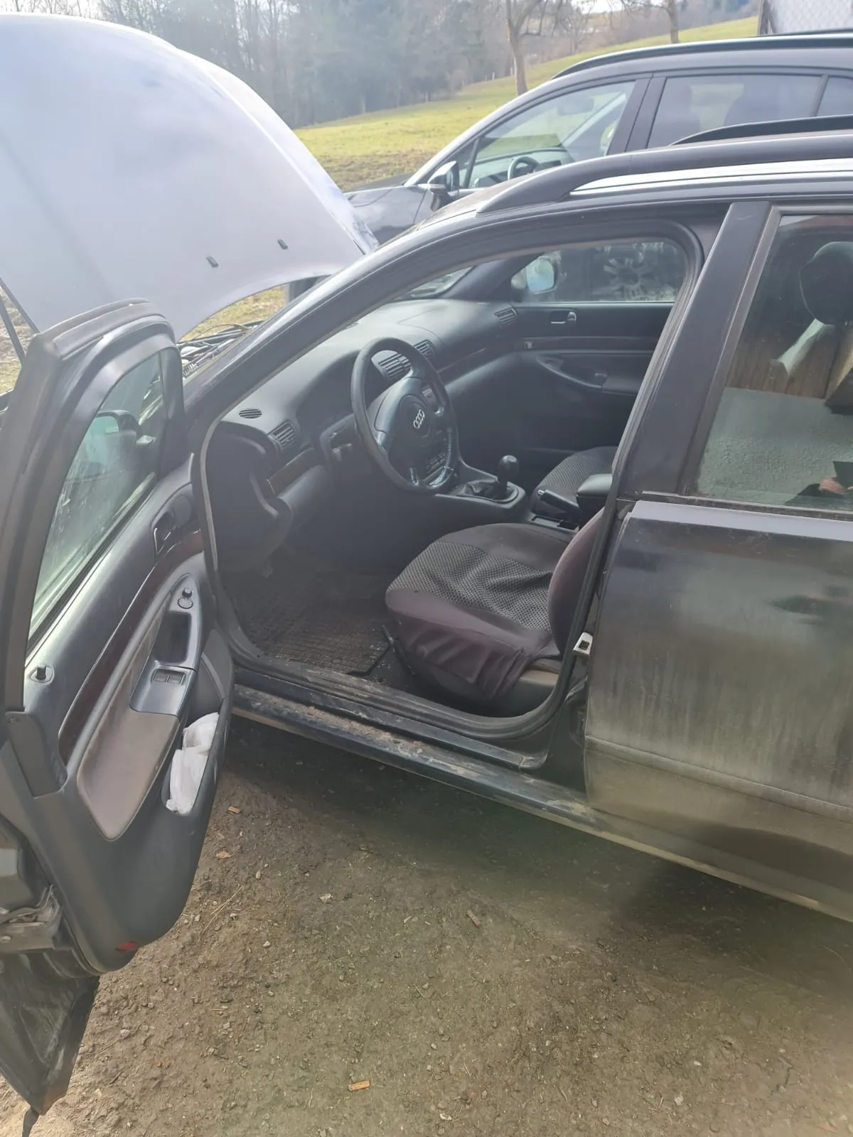 Ciemnoszary samochód marki Audi z otwartą maską i otwartymi drzwiami kierowcy. Wnętrze z widoczną kierownicą z logo Audi, dźwignią zmiany biegów i siedzeniami. W tle drugi samochód, trawa i drzewa. Samochód stoi na brudnej, ziemistej powierzchni.