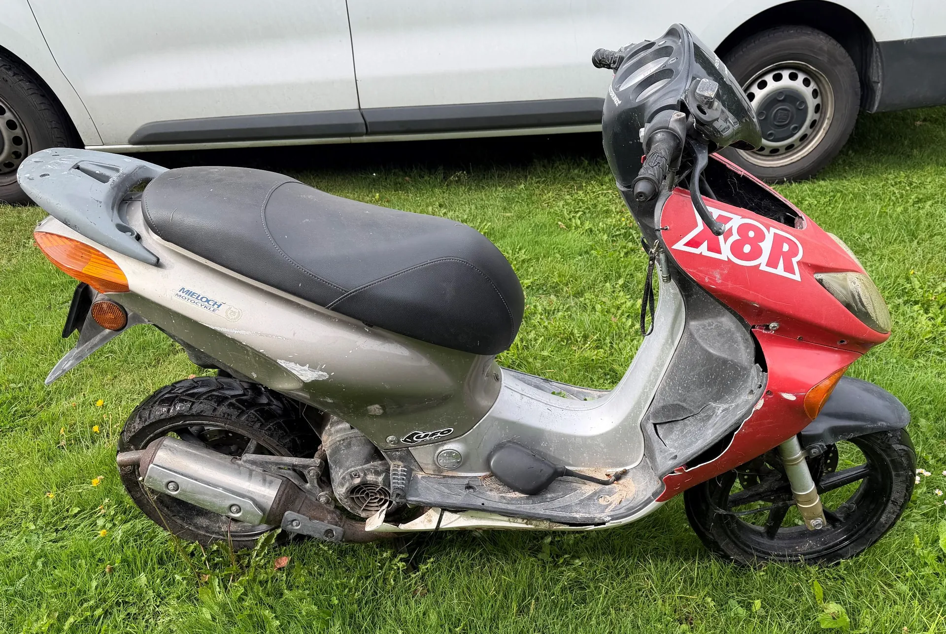 Srebrno-czerwony skuter marki Aprilia z napisem 'X8R' na przedniej owiewce, zaparkowany na zielonej trawie na tle białego samochodu. Pojazd jest w złym stanie technicznym, z widocznymi uszkodzeniami plastikowych elementów nadwozia w dolnej części oraz zabrudzeniami.