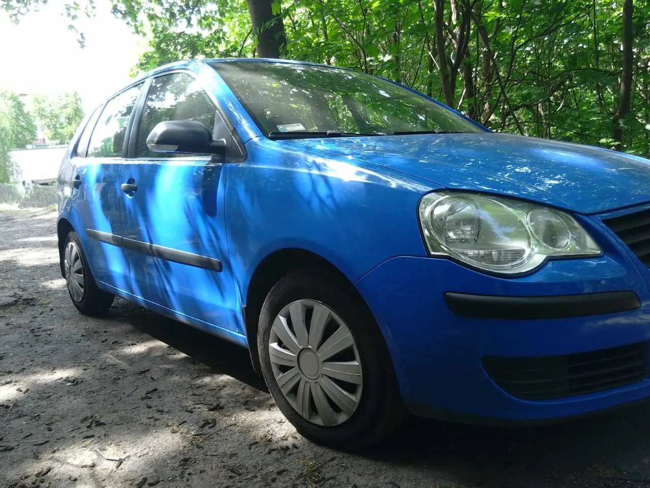 Niebieski samochód osobowy typu hatchback zaparkowany na drodze z żwiru. W tle zielone drzewa i krzewy. Samochód ma srebrne felgi, czarne zderzaki i lusterka boczne. Słońce oświetla pojazd, rzucając cienie na ziemię.