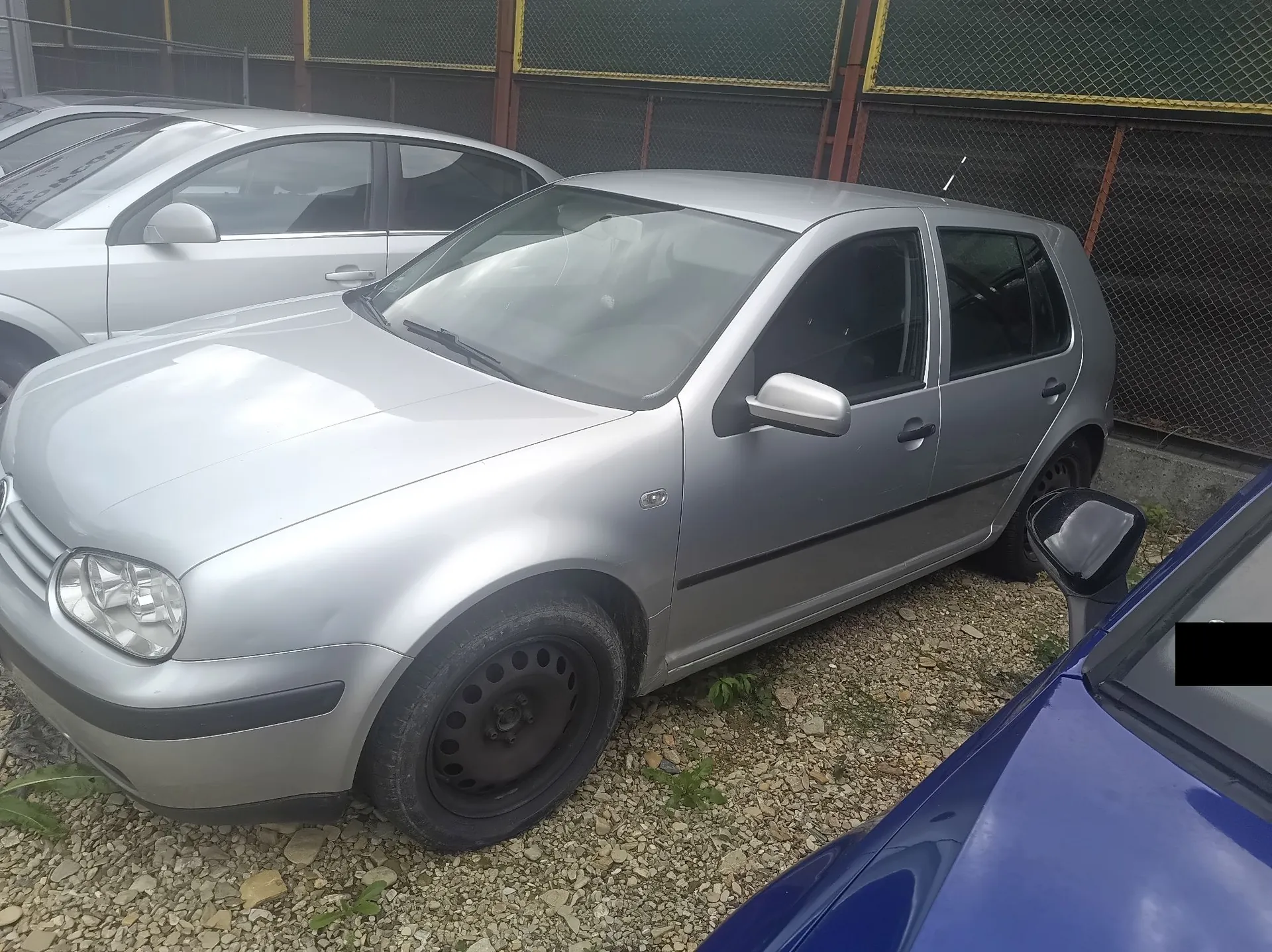 Srebrny samochód osobowy marki Volkswagen Golf zaparkowany na żwirowym placu. Pojazd jest widoczny z przodu i z boku, posiada czarne felgi stalowe. W tle widoczny jest fragment innego srebrnego samochodu oraz ciemny płot z siatką. Po prawej stronie kadru widoczny jest fragment niebieskiego samochodu.