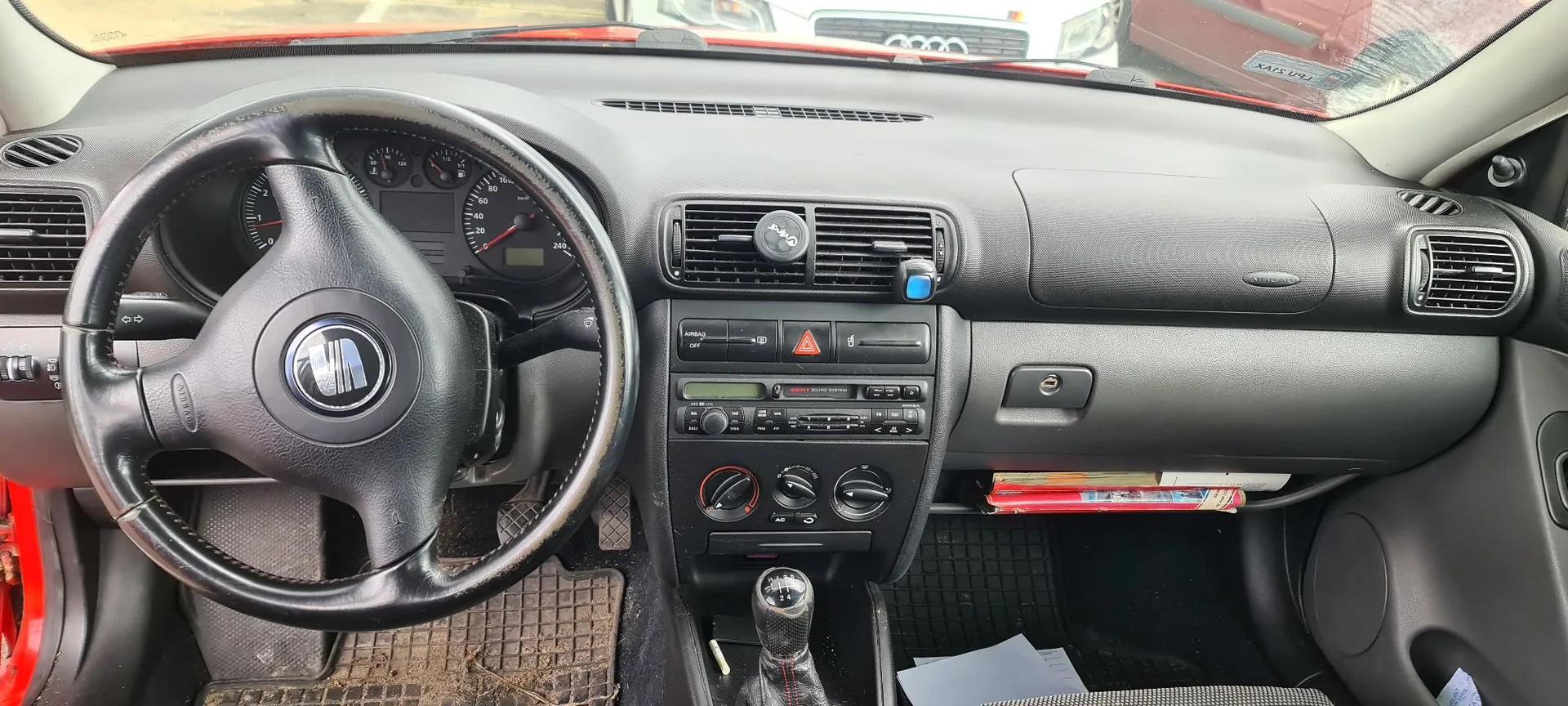 Wnętrze samochodu marki SEAT, widok z perspektywy kierowcy na deskę rozdzielczą. Po lewej stronie znajduje się czarna, skórzana kierownica z logo SEAT w centrum. Za kierownicą widoczne są analogowe wskaźniki prędkości i obrotów. W centralnej części deski znajduje się system wentylacji z kratkami nawiewu, radio z wyświetlaczem oraz pokrętła sterowania klimatyzacją. Po prawej stronie znajduje się schowek dla pasażera. W dolnej części widoczna jest wajcha zmiany biegów oraz gumowe dywaniki na podłodze.