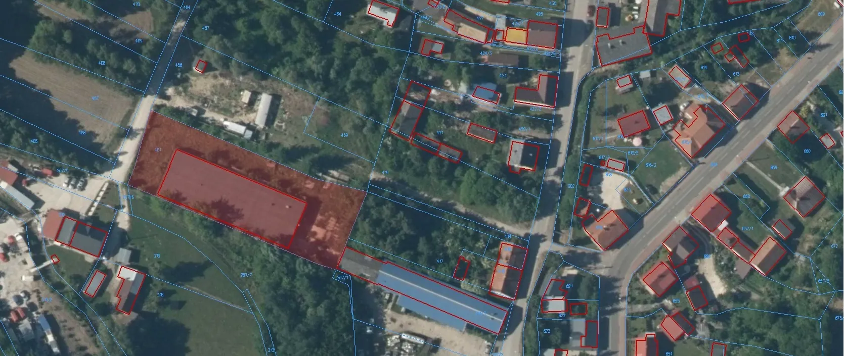 Zdjęcie lotnicze terenu zabudowanego i niezabudowanego z nałożoną siatką geodezyjną. Na obrazie widoczne są granice działek zaznaczone niebieskimi liniami oraz obrysy budynków zaznaczone czerwonymi liniami. Część działek i budynków jest wypełniona półprzezroczystym czerwonym kolorem. Widoczne są domy jednorodzinne, drogi oraz obszary zielone z drzewami i krzewami. Na działkach znajdują się numery ewidencyjne, np. 451, 450, 417.