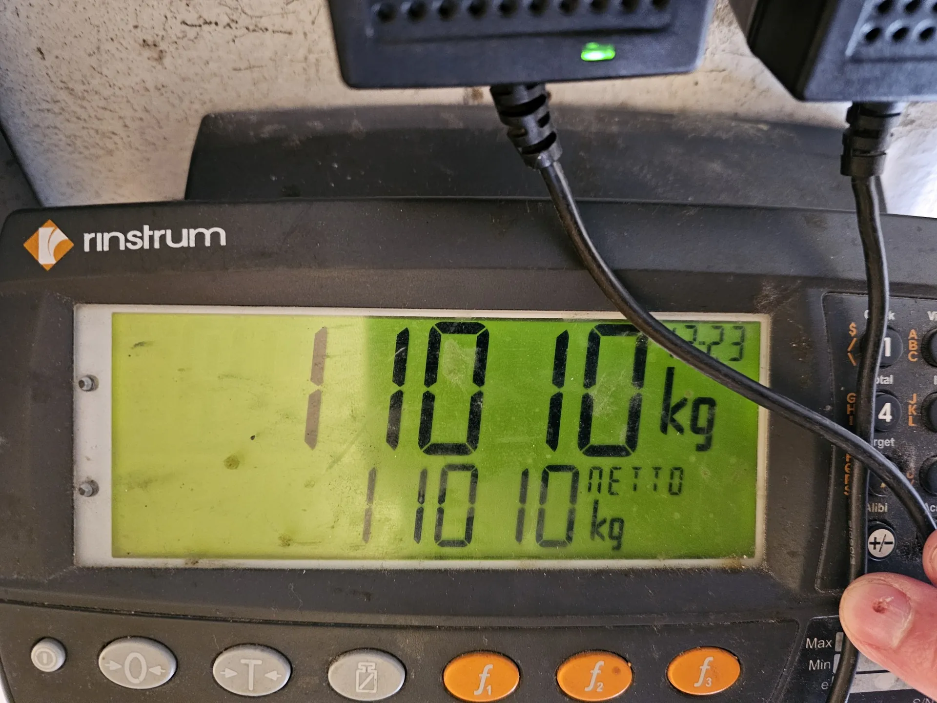 Zbliżenie na cyfrowy wyświetlacz wagi marki rinstrum. Na ekranie widnieje liczba 110.10 kg, a poniżej mniejszym drukiem powtórzona wartość 110.10 kg oraz napis NETTO. Poniżej ekranu znajdują się przyciski sterujące, w tym trzy pomarańczowe przyciski z oznaczeniami f1, f2 i f3. Do urządzenia podłączone są czarne kable.
