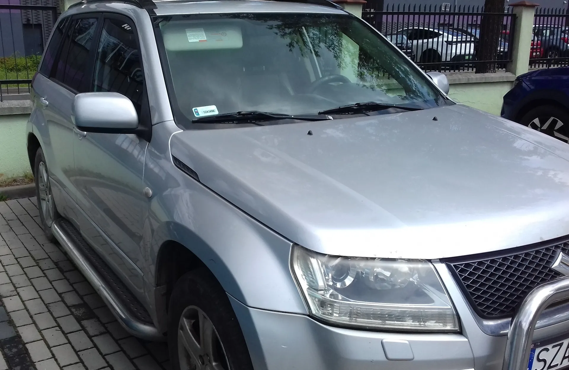 Srebrny samochód typu SUV zaparkowany na kostce brukowej. Widoczny jest przód i lewy bok pojazdu, w tym reflektor, lusterko boczne oraz stopień boczny. W tle znajduje się ogrodzenie i fragment innego białego samochodu.