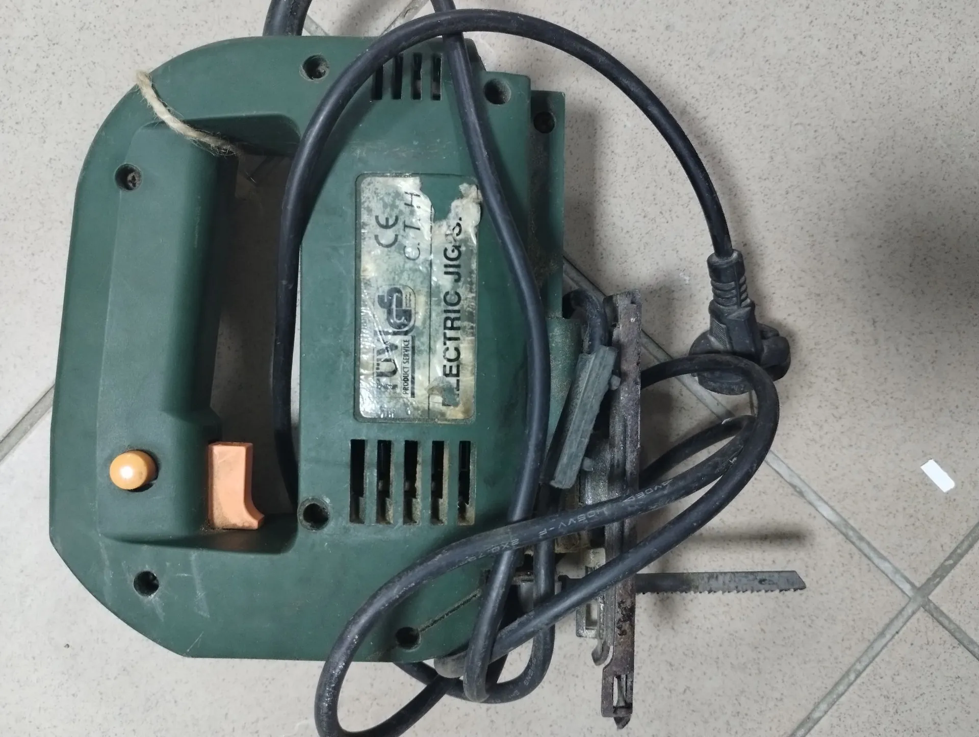 Zielona wyrzynarka elektryczna leżąca na jasnych kafelkach. Urządzenie ma czarny kabel zasilający owinięty wokół obudowy. Na boku widoczna jest metalowa tabliczka znamionowa z napisem 'ELECTRIC JIGS.', znakiem CE oraz innymi oznaczeniami technicznymi. Widoczny jest pomarańczowy przełącznik oraz brązowy element plastikowy.