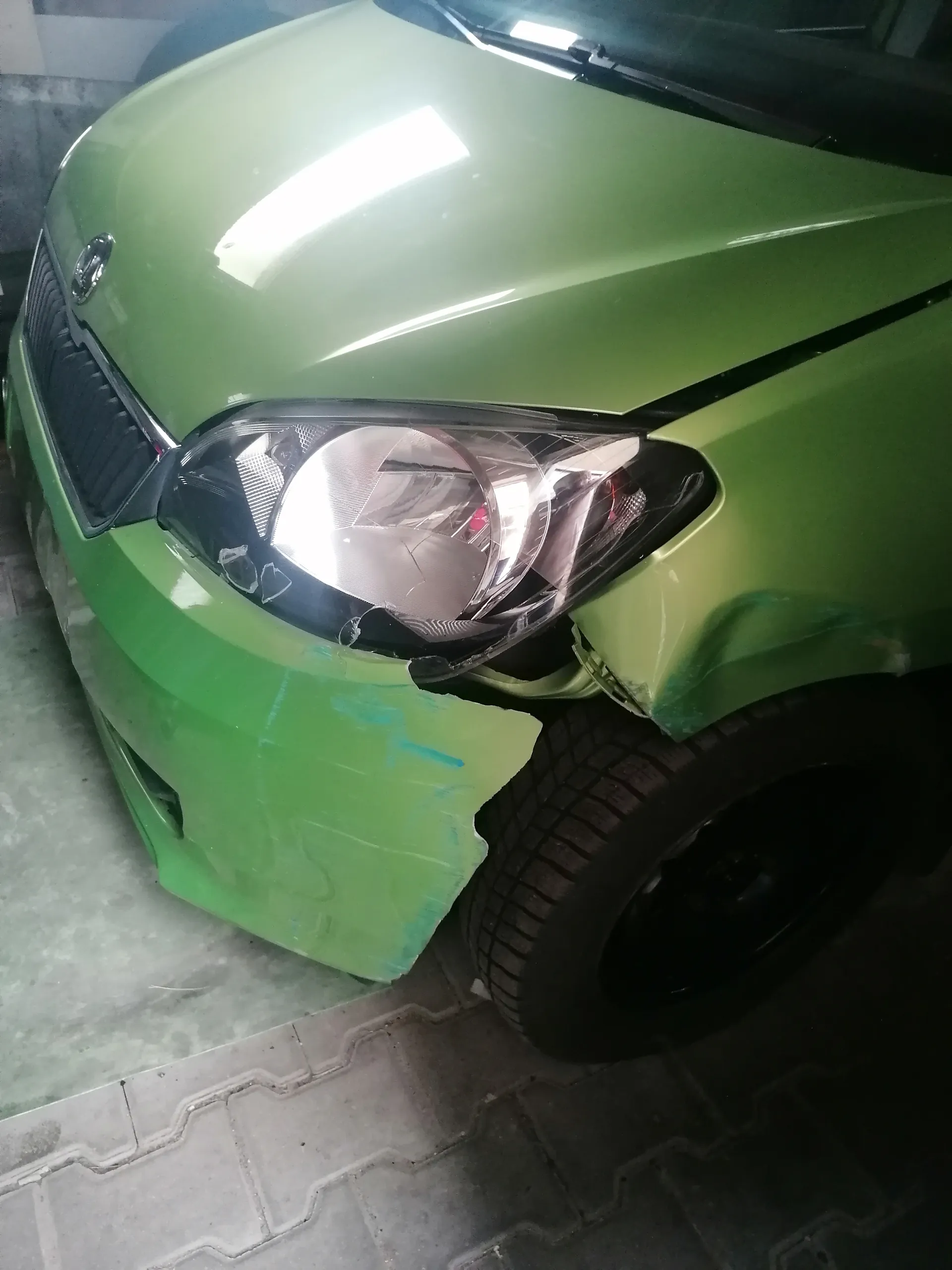 Zielony samochód marki Skoda z uszkodzonym przednim lewym reflektorem, który ma pęknięcia i częściowo usuniętą osłonę. Widoczna część pasa przedniego z uszkodzonym zderzakiem. Samochód zaparkowany na brukowanej powierzchni, prawdopodobnie w garażu. Na masce widoczne odbicia światła.