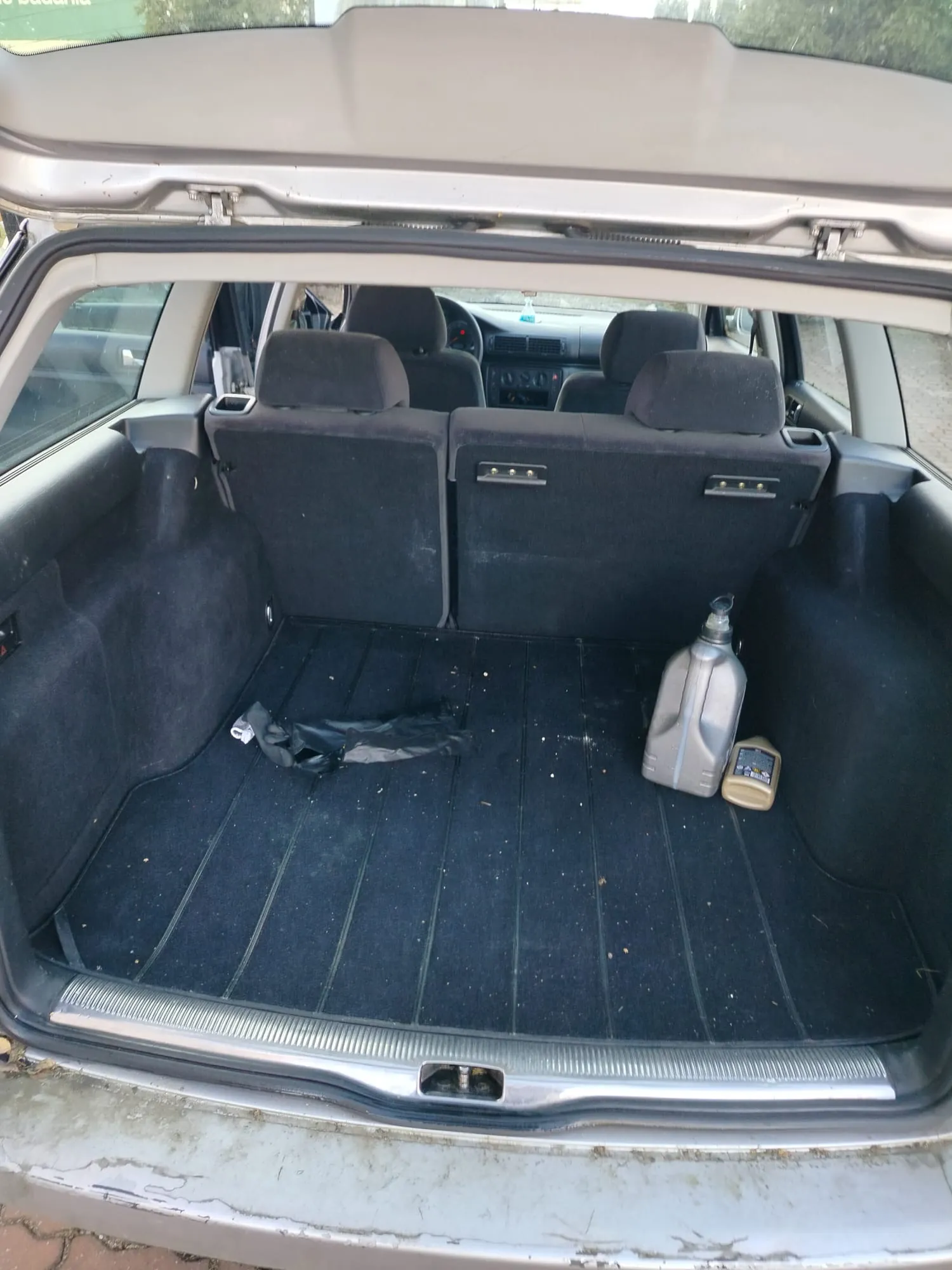 Otwarty bagażnik samochodu z ciemnym wykładziną. Wewnątrz znajdują się: duża srebrna kanistra, mała brązowa butelka z nieczytelnym napisem oraz czarny plastikowy przedmiot. Widoczne są złożone tylnie siedzenia, a przez przednią szybę widać fragment kabiny z kierownicą i deską rozdzielczą. Samochód ma jasnoszary kolor, bagażnik jest otwarty, a na zewnątrz widoczny jest bruk i zieleń.