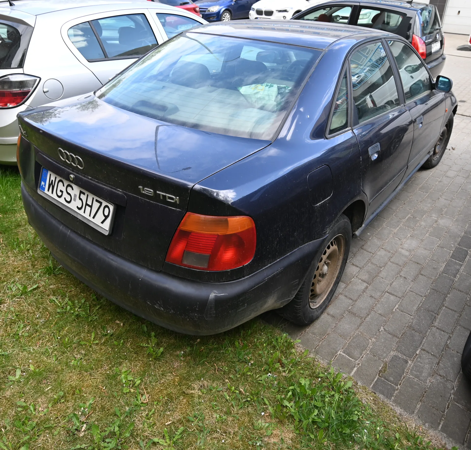 Czarny samochód marki Audi z widocznym tyłem, na którym znajduje się tablica rejestracyjna PL WGS 5H79 oraz napis 1.9 TDI. Samochód zaparkowany jest na brukowanej powierzchni obok trawnika. W tle widoczne są inne pojazdy: srebrny i czarny samochód. Na szybie tylnej widoczne są odbicia oraz białe przedmioty wewnątrz pojazdu.