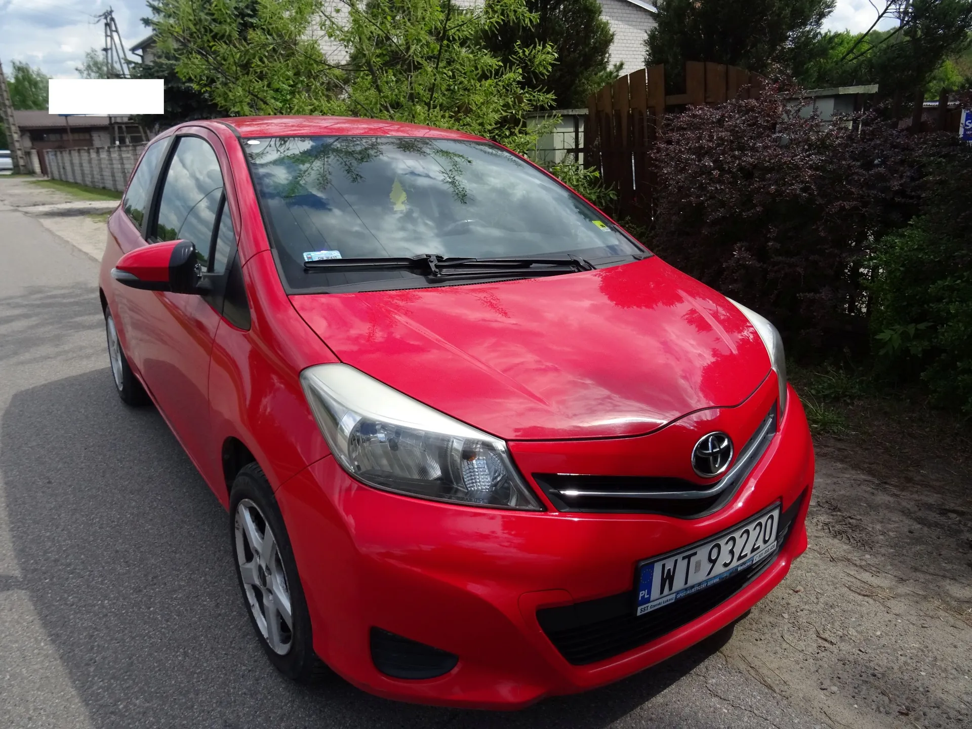 Czerwony samochód osobowy marki Toyota zaparkowany na asfaltowej drodze. Pojazd jest widoczny z przodu i z boku. Na samochodzie znajduje się polska tablica rejestracyjna o numerze WT 93220. W tle widoczne są zielone krzewy, drzewa oraz fragment ogrodzenia.