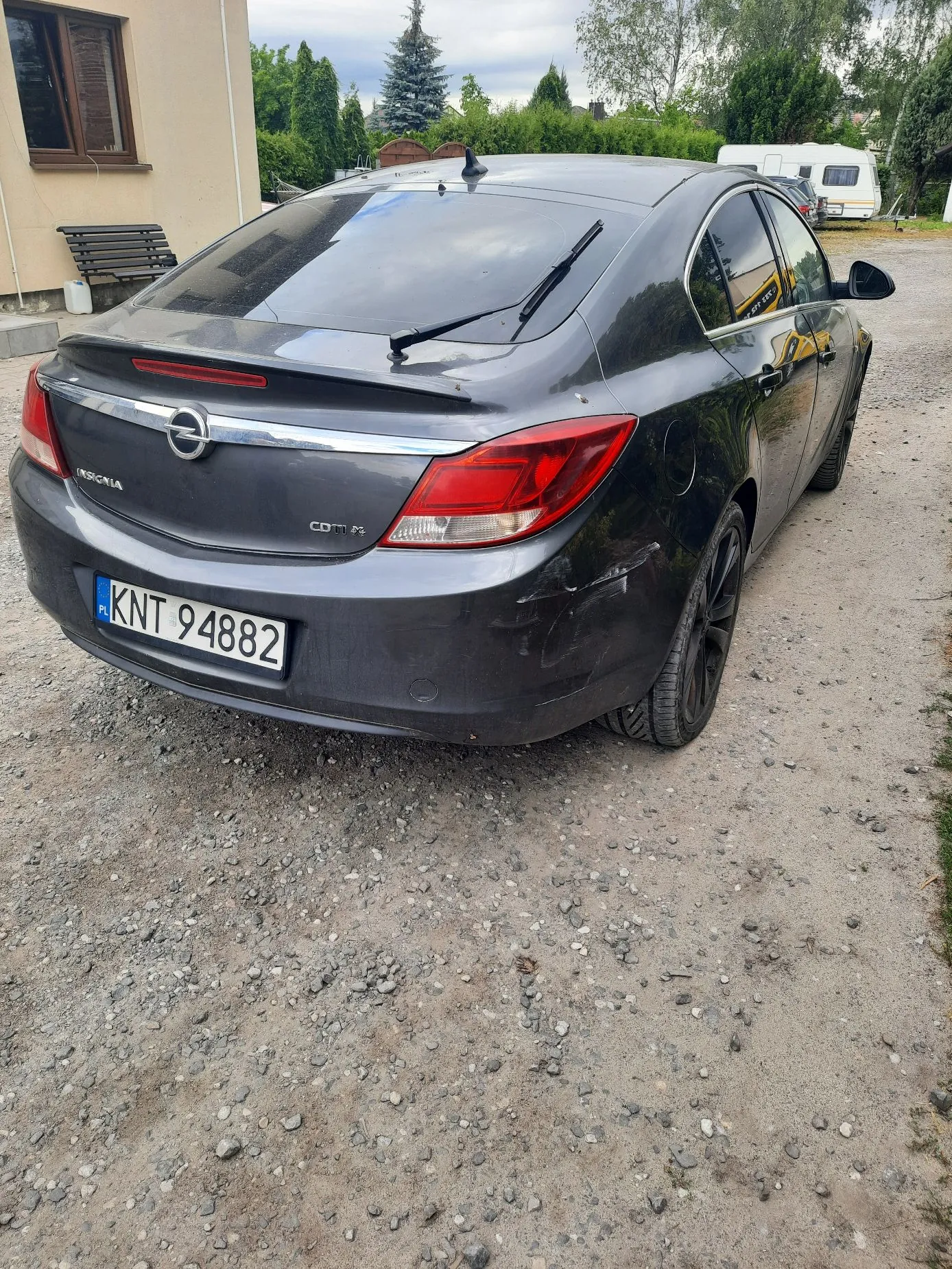 Tylny widok ciemnoszarego samochodu marki Opel Insignia zaparkowanego na żwirowym podjeździe. Na klapie bagażnika widoczne są napisy 'INSIGNIA' oraz 'CDTI'. Samochód ma polską tablicę rejestracyjną o numerze KNT 94882. W tle widać fragment jasnego budynku z oknem, zielone drzewa oraz białą przyczepę kempingową.