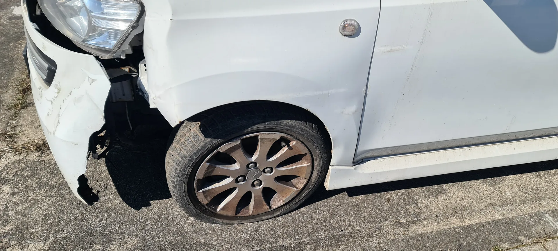 Biały samochód z widocznymi uszkodzeniami na przedniej lewej stronie. Zniszczony zderzak, pęknięty i częściowo odłączony, odsłaniający wewnętrzne elementy. Widoczne przednie lewe koło z metalową felgą o brązowym odcieniu i oponą. Samochód stoi na betonowej powierzchni z fragmentami trawy po lewej stronie. Część przedniego reflektora i kierunkowskazu jest widoczna.