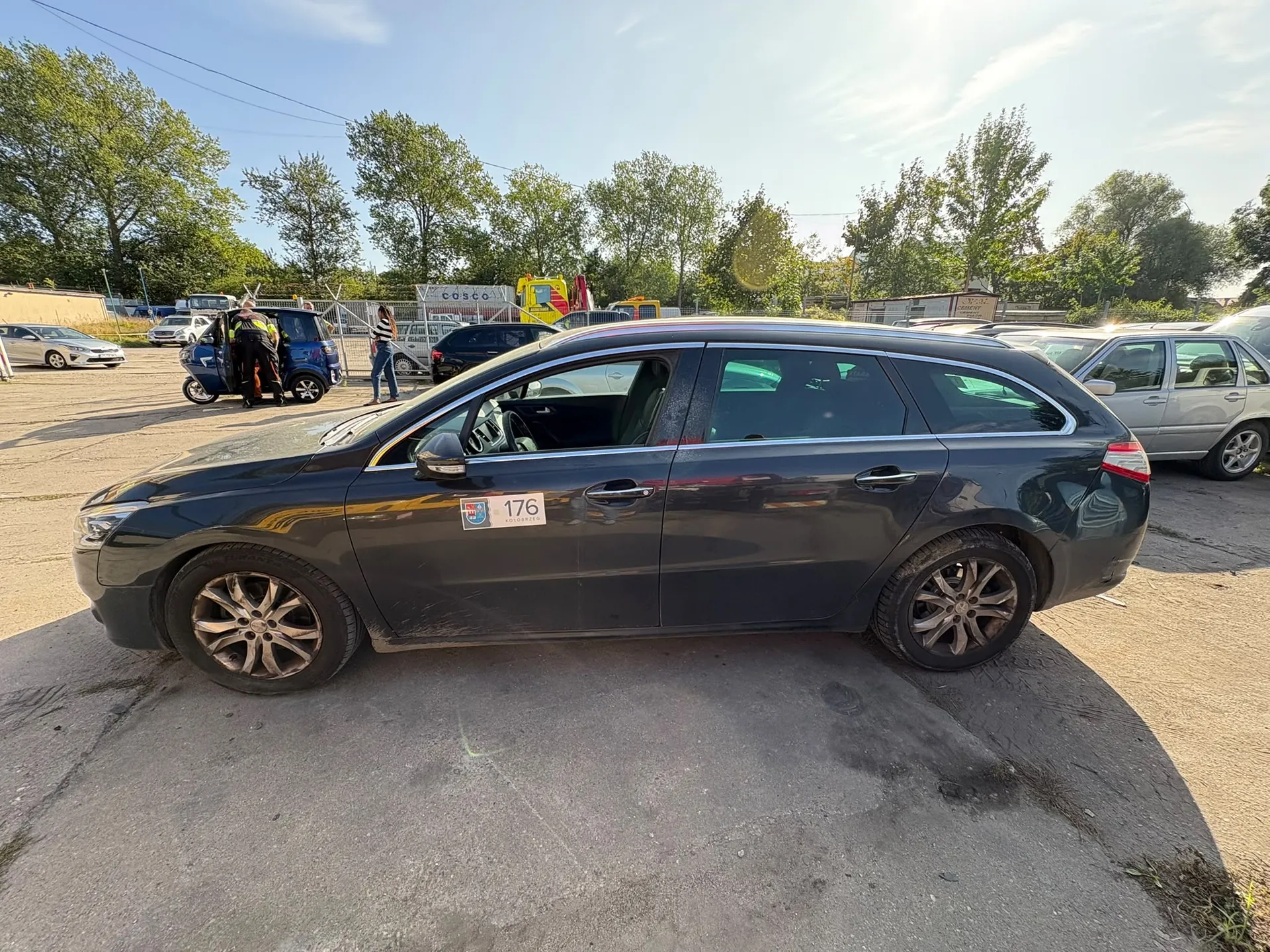 Czarny samochód typu kombi zaparkowany bokiem na asfaltowym placu. Na przednich drzwiach kierowcy znajduje się naklejka z numerem 176 i logo. W tle widoczne są inne zaparkowane samochody, w tym niebieski pojazd przy którym stoi osoba w odblaskowej kamizelce, oraz zielone drzewa pod jasnym, słonecznym niebem.