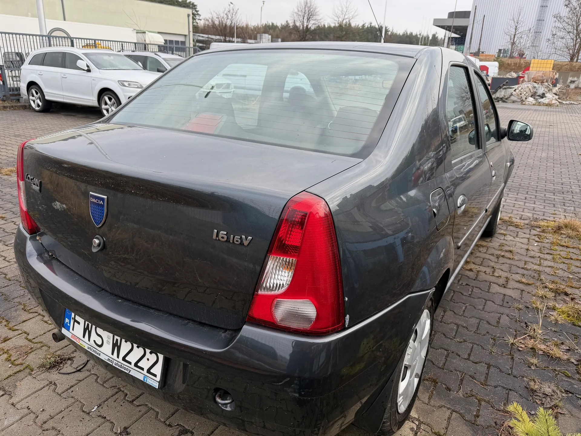 Czarny samochód marki Dacia Logan z widocznym tyłem, na którym znajduje się logo Dacia, napis 1.6 16V oraz tablica rejestracyjna PL FWS W222. Samochód stoi na brukowanej powierzchni, w tle widoczne są inne pojazdy, budynek, ogrodzenie i drzewa. Na tablicy rejestracyjnej widoczny jest dodatkowy napis Brico Wschowa ...mój ulubiony.