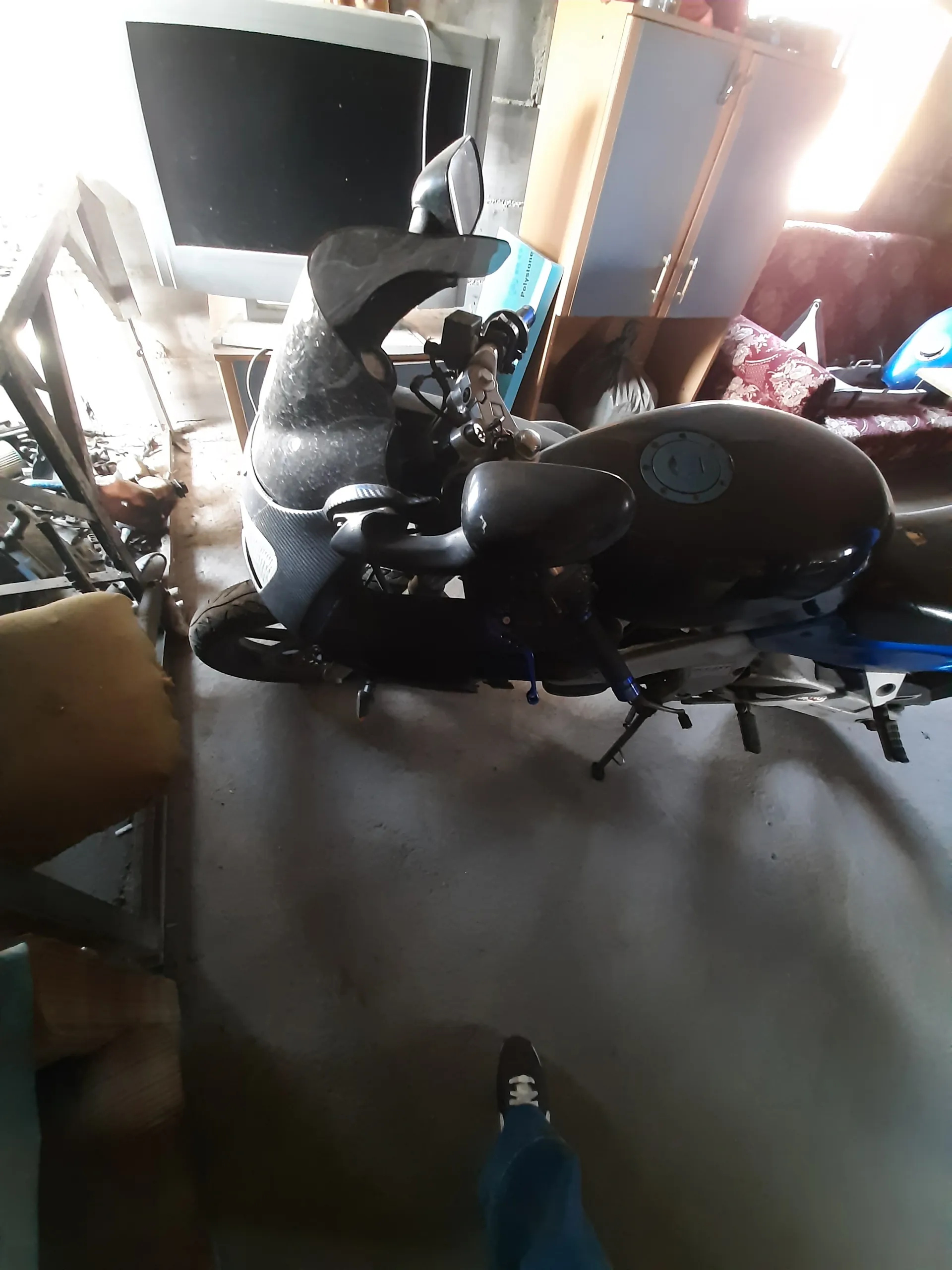 Ciemny motocykl typu skuter z niebieskimi akcentami na betonowej podłodze. W tle szafka z szufladami, telewizor lub monitor, narzędzia oraz czerwona poduszka z wzorem. Widoczna noga osoby w niebieskich dżinsach i czarnym butie z białymi sznurówkami po prawej stronie. Oświetlenie pochodzi z lewego górnego rogu.