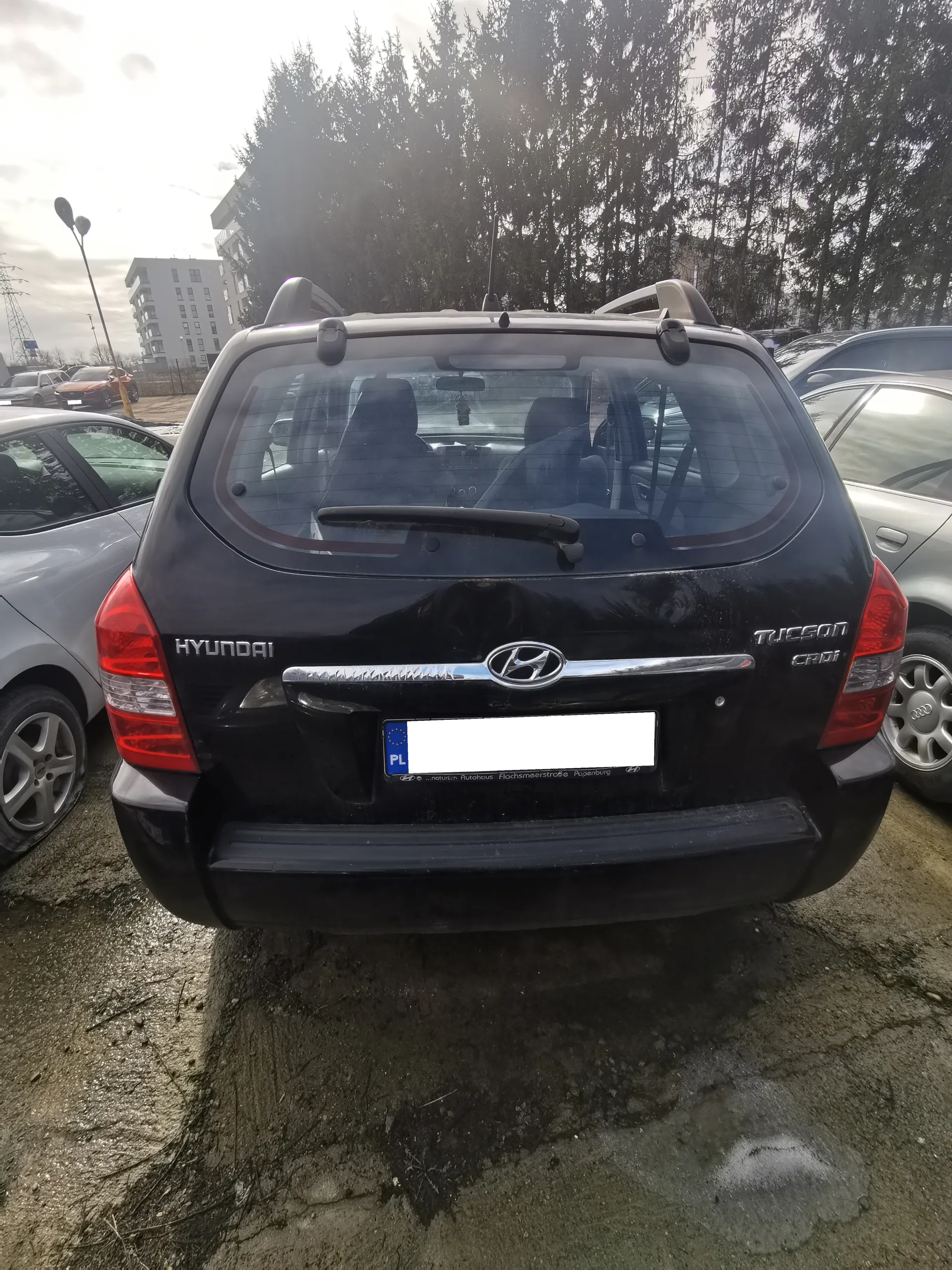 Tył czarnego samochodu Hyundai Tucson CRDi zaparkowanego na otwartym parkingu. Widoczne są czerwone światła tylnego, logo marki, napisy 'HYUNDAI' po lewej stronie i 'TUCSON CRDi' po prawej stronie. Tablica rejestracyjna z niebieskim pasem z napisem 'PL' i białym polem z zamazanym numerem. W tle inne zaparkowane samochody, wielopiętrowy budynek, drzewa oraz pochmurne niebo. Podłoga parkingu jest brudna z widocznymi plamami i szczątkami roślin.