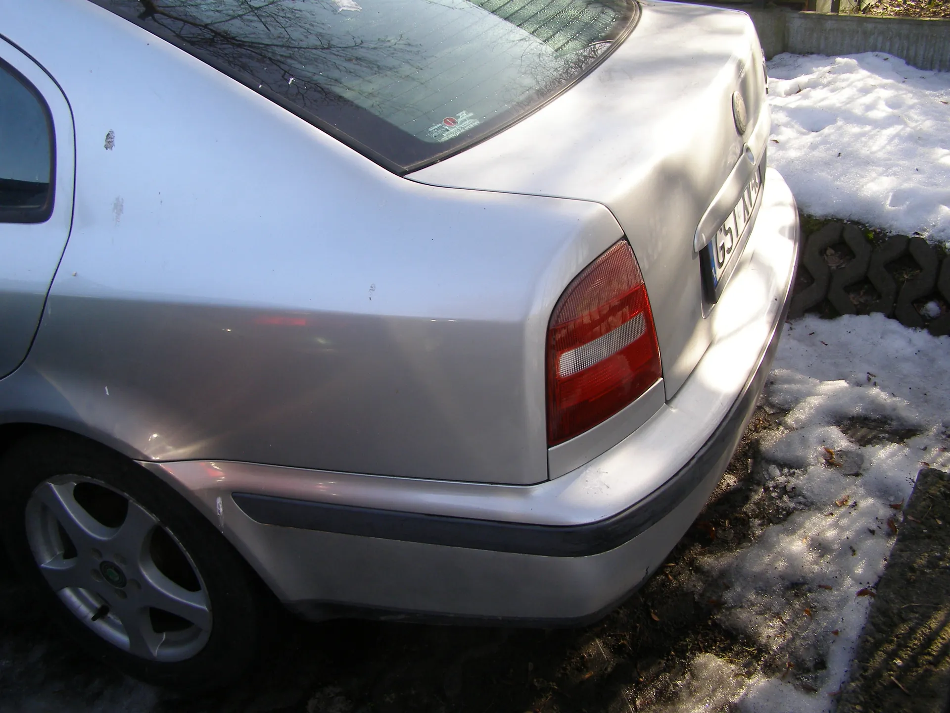 Tylne widok srebrnego samochodu z czerwonym światłem tylnym i tablicą rejestracyjną z widocznymi znakami G51. Na tle śnieg na ziemi, kamienna ściana oraz odbicie drzew w tylnej szybie. Samochód ma widoczne ślady na karoserii.