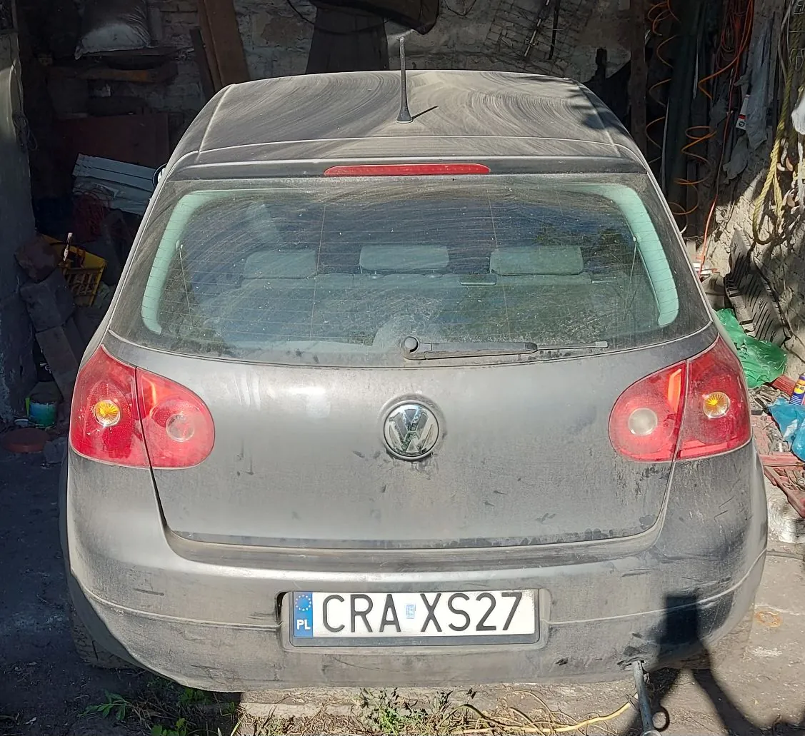 Widok z tyłu na zakurzony, ciemnoszary samochód marki Volkswagen. Pojazd posiada tablicę rejestracyjną o numerze CRA XS27 z oznaczeniem PL. Samochód stoi w pomieszczeniu przypominającym garaż lub warsztat.