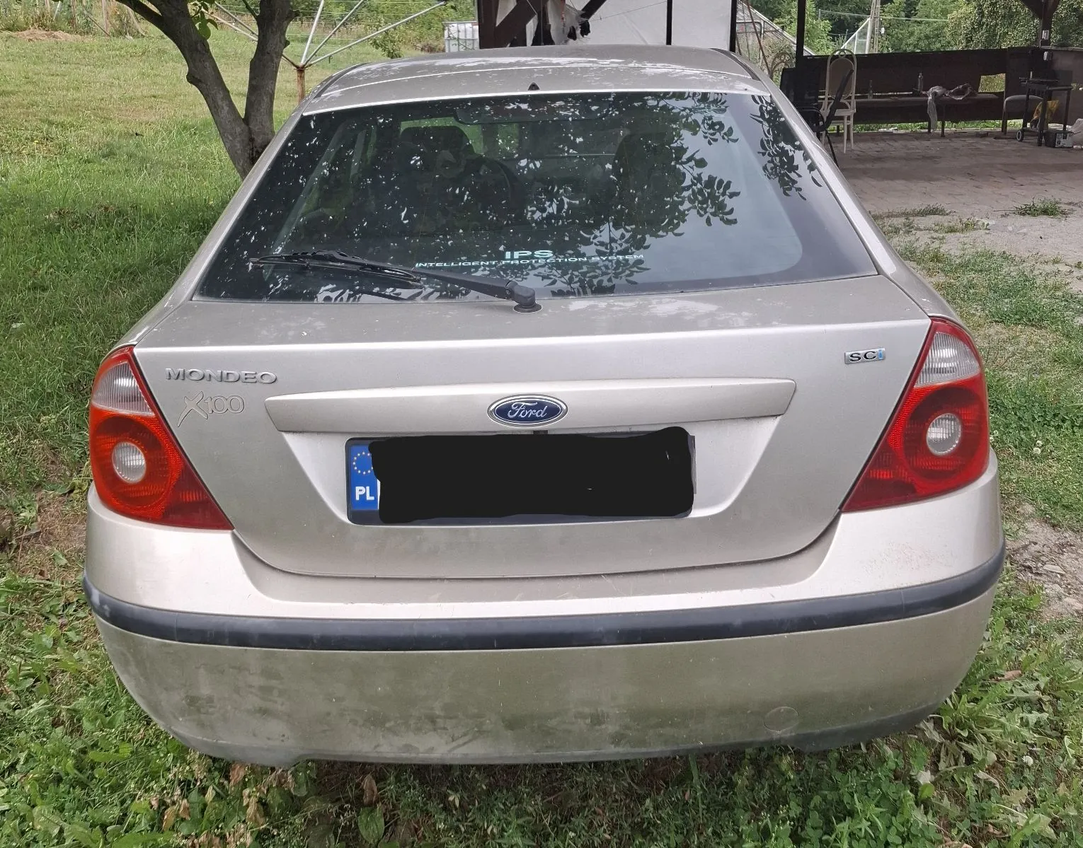 Tylny widok srebrnego samochodu marki Ford Mondeo SC. Na klapie bagażnika widoczne są napisy 'MONDEO', '1.8 TDCi' oraz logo 'Ford'. Samochód posiada polską tablicę rejestracyjną z symbolem PL, której numery są zakryte czarnym polem. Pojazd stoi na trawiastej powierzchni.
