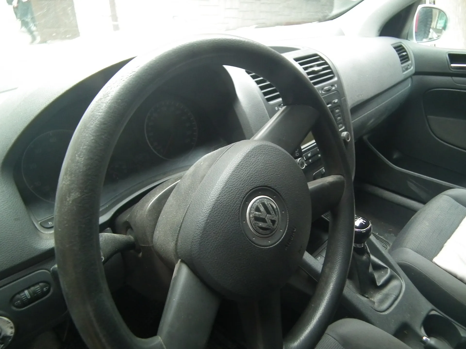 Wnętrze samochodu z widocznym kierownicą marki Volkswagen, tablicą przyrządów z licznikami prędkości, dźwignią zmiany biegów, czarnymi siedzeniami oraz fragmentem deski rozdzielczej z wentylacjami i przyciskami. W tle widoczne jest przednie szyby i lusterko boczne.