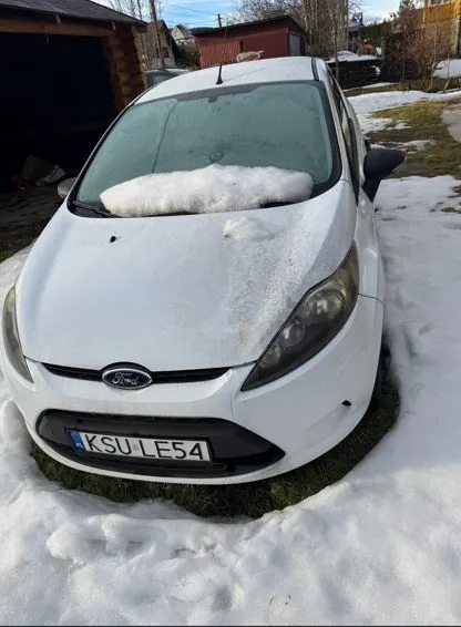 Biały samochód marki Ford pokryty śniegiem, zaparkowany na śnieżnej powierzchni z widoczną trawą. Na masce i przedniej szybie leży warstwa śniegu. Tablica rejestracyjna z napisem K SU LE54. W tle drewniany garaż, czerwony budynek, drzewa i ogrodzenie. Niebo jest jasne.