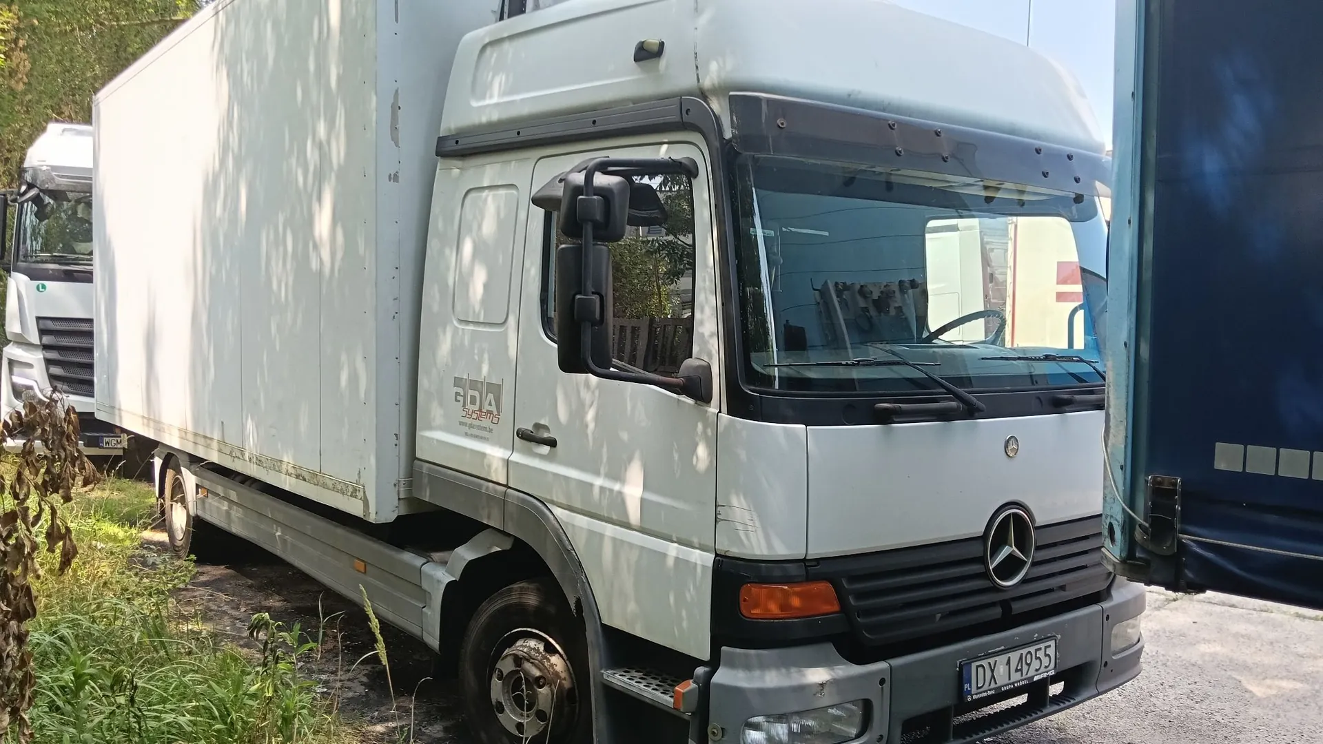 Biała ciężarówka marki Mercedes-Benz z zabudową furgonową, zaparkowana na zewnątrz. Na drzwiach kabiny widnieje napis 'GDA'. Pojazd posiada tablicę rejestracyjną o numerze DX 14955.