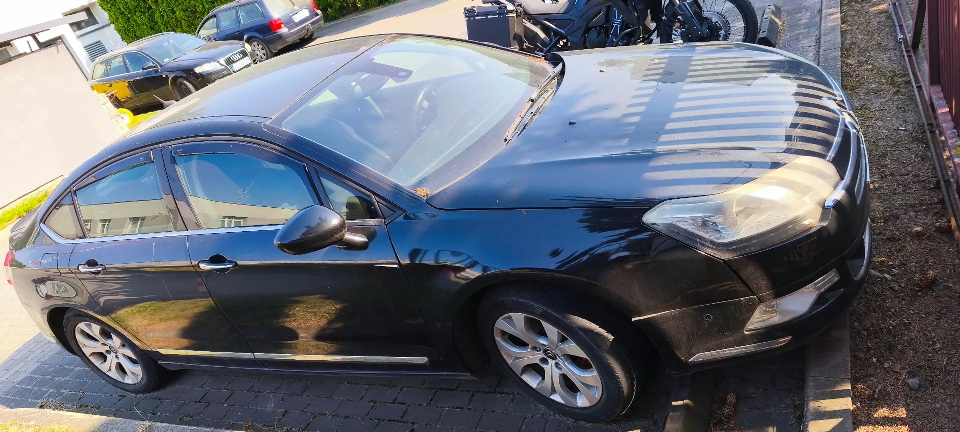 Czarny samochód osobowy zaparkowany na kostce brukowej, widoczny z perspektywy przedniego lewego boku. W tle widać inne zaparkowane samochody oraz fragmenty zabudowań i zieleni.