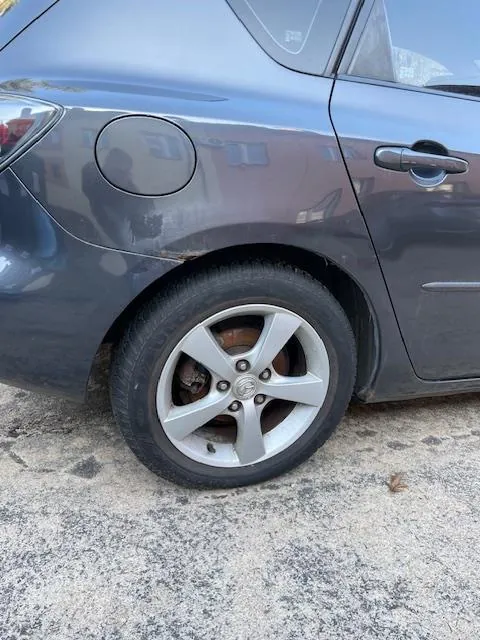 Zbliżenie na tylną część ciemnoszarego samochodu, obejmujące koło z srebrnym felgiem, oponę oraz fragment nadwozia z pokrywą zbiornika paliwa. Samochód stoi na betonowej powierzchni z widocznymi pęknięciami i małym liściem. W tle odbicia na nadwoziu sugerują obecność budynków lub innych pojazdów.