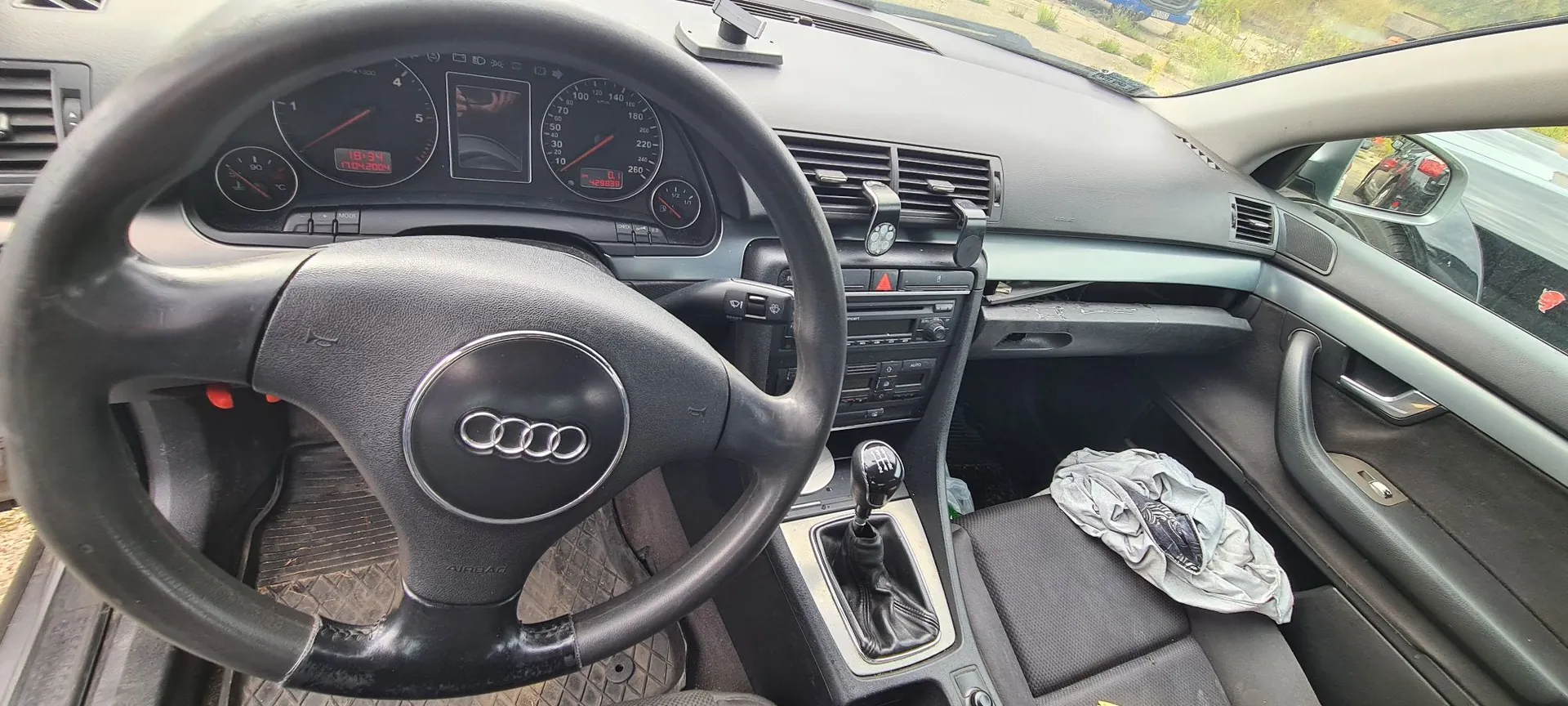 Wnętrze samochodu marki Audi widziane z perspektywy kierowcy. Widoczna jest czarna kierownica z logo Audi w centrum, deska rozdzielcza z analogowymi wskaźnikami prędkości i obrotomierzem, konsola środkowa z radiem i lewarkiem zmiany biegów oraz przedni fotel pasażera, na którym leży szara tkanina.