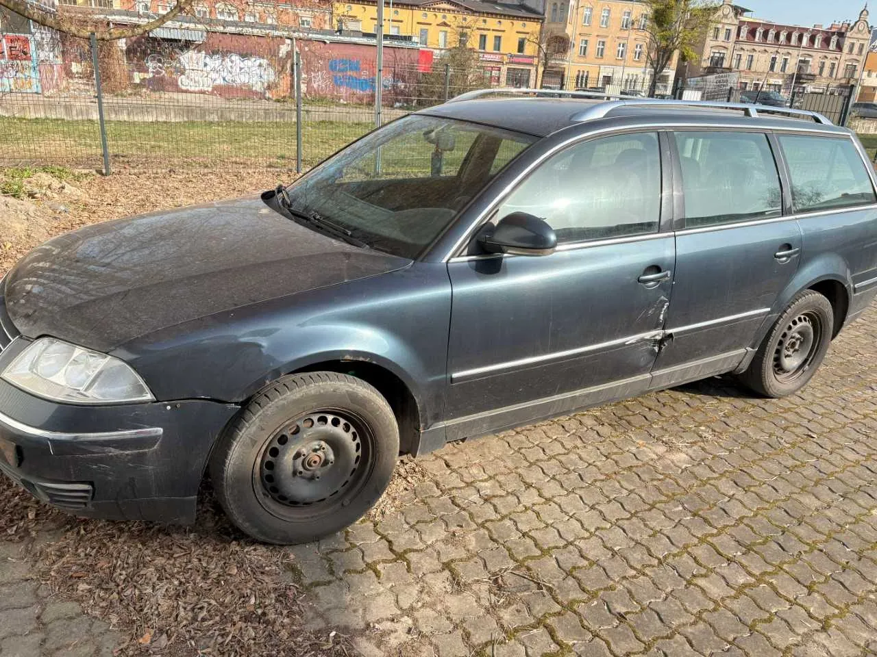 Ciemnoszary samochód typu kombi zaparkowany na brukowanej powierzchni. Pojazd ma uszkodzony przód, brakującą pokrywę felgi na przednim lewym kole oraz widoczne zadrapania na boku. W tle metalowe ogrodzenie, trawa z suchymi liśćmi, budynki z kolorowymi graffiti na ścianach oraz drzewa. Na pierwszym planie suchy liść i brud na powierzchni samochodu.