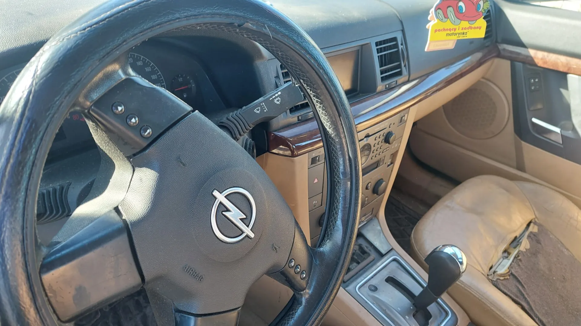Wnętrze samochodu z kierownicą marki Opel, na której widoczny jest logo i napis 'AIRBAG'. W tle zegary samochodowe, w tym prędkościomierz, oraz deska rozdzielcza z drewnianą obwódką. Po prawej stronie widoczne są beżowe, zużyte siedzenia z uszkodzonym materiałem. Na desce rozdzielczej naklejka z napisem 'pachnący i zadbaný motorynka' oraz ilustracją czerwonego samochodu. W centrum konsoli centralnej znajduje się dźwignia zmiany biegów.