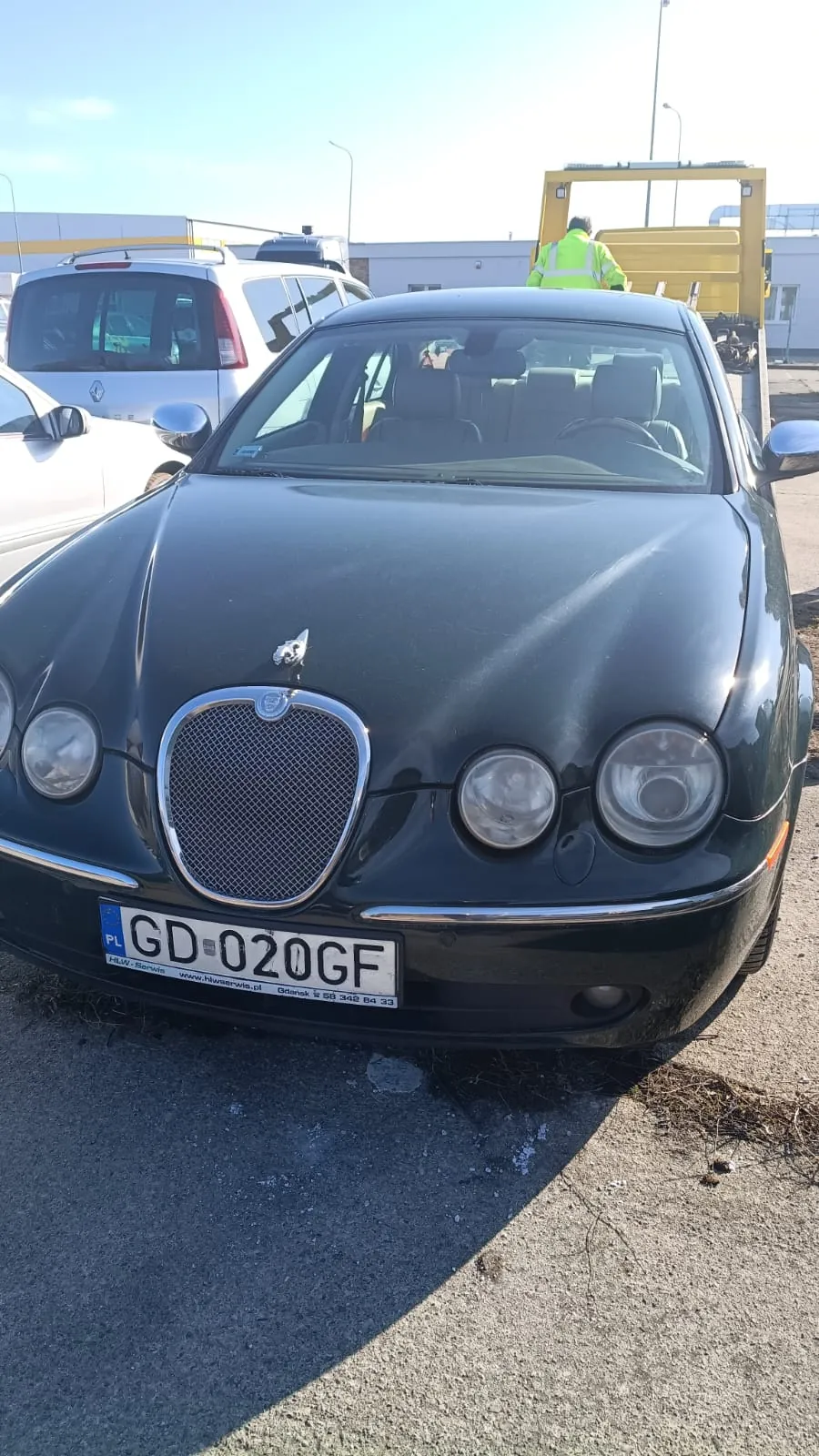 Ciemnozielony samochód marki Jaguar z przodu, z widoczną tablicą rejestracyjną PL GD-020GF oraz napisem www.hiwservis.pl i numerem telefonu pod nią. W tle białe samochody, żółty pojazd z osobą w jasnozielonej kamizelce odblaskowej oraz budynki przemysłowe. Scena na otwartym placu asfaltowym w słoneczny dzień.