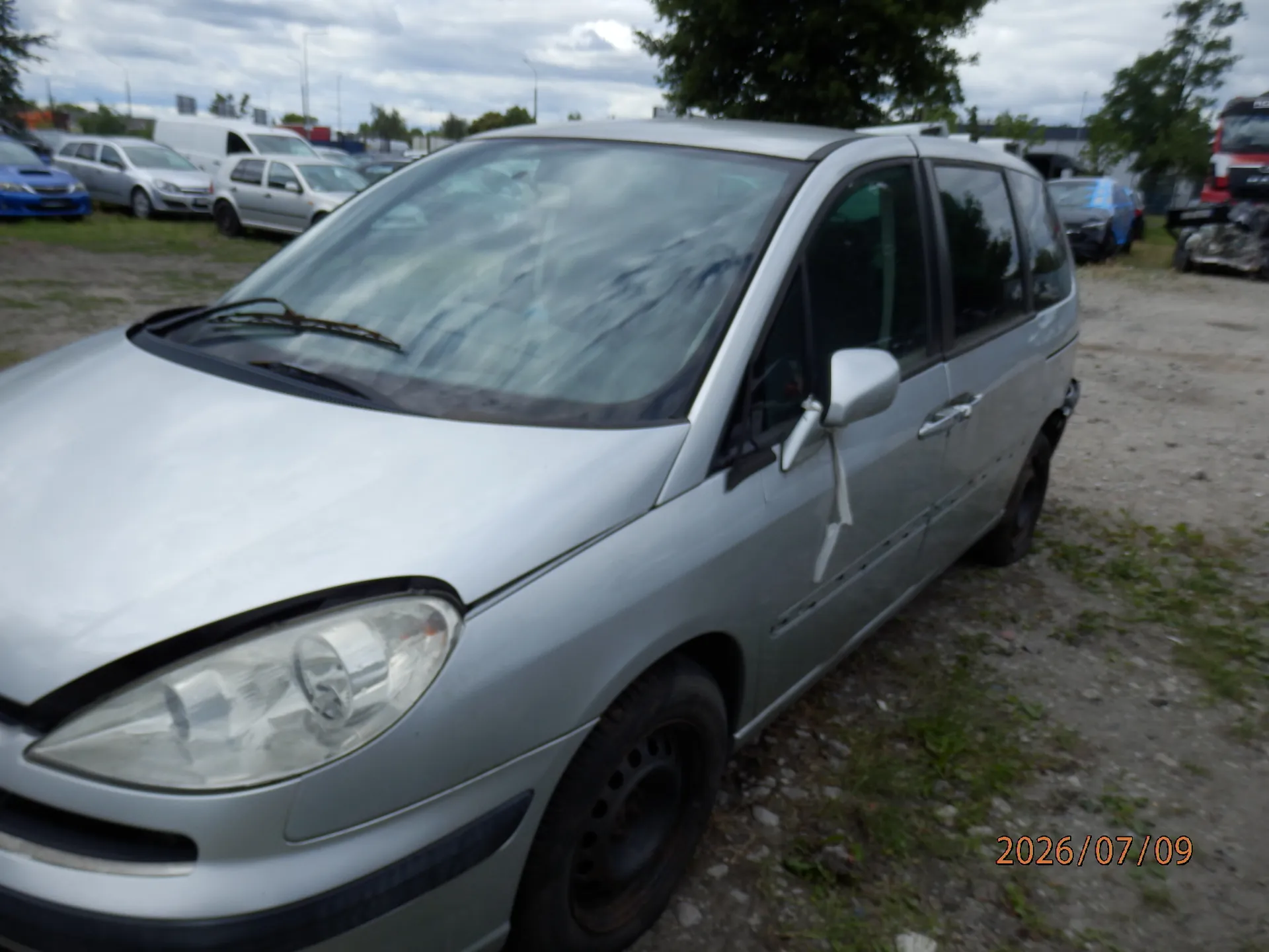 Srebrny samochód typu minivan zaparkowany na nieutwardzonym placu. Widok z przodu i z boku pojazdu. W tle widoczne są inne samochody oraz drzewa pod zachmurzonym niebem. W prawym dolnym rogu znajduje się pomarańczowy napis z datą: 2026/07/09.