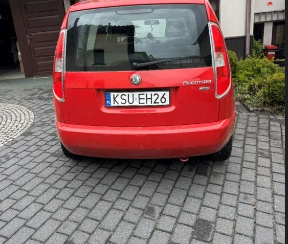 Czerwony samochód Škoda Roomster widziany od tyłu, z tablicą rejestracyjną KSU EH26. Pojazd stoi na brukowanej powierzchni. W tle widoczne są brama garażowa, krzewy i fragment budynku.