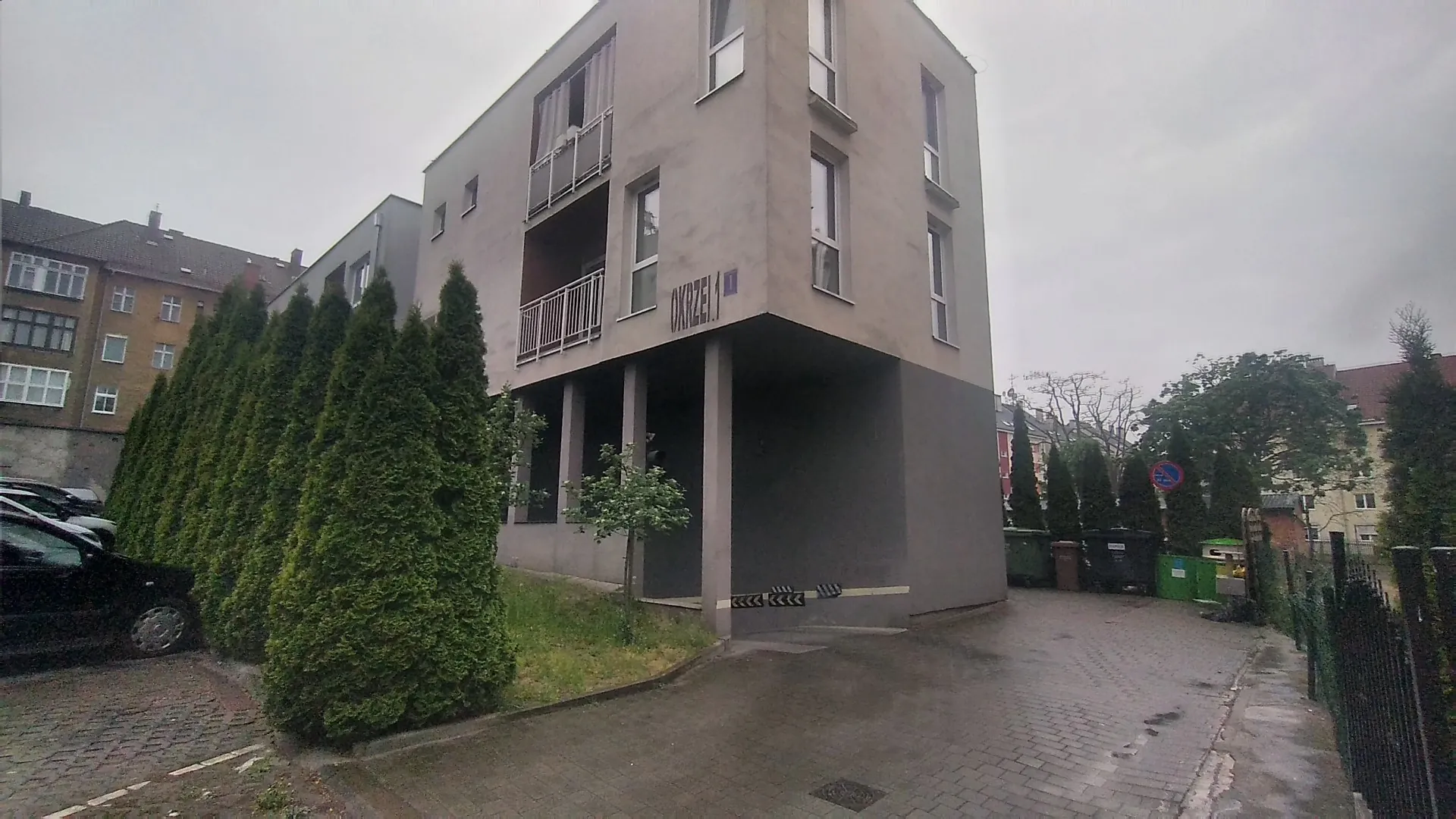 Nowoczesny, szary, wielorodzinny budynek mieszkalny z balkonami i dużymi oknami. Na elewacji widoczny jest napis 'OKRZEI 1'. Przed budynkiem rośnie rząd wysokich, gęstych tuj oraz mniejsze drzewko. Po lewej stronie zaparkowany jest czarny samochód. Po prawej stronie znajduje się podjazd z kostki brukowej prowadzący do zaplecza z kolorowymi kontenerami na odpady. W tle widoczne są inne budynki i drzewa pod zachmurzonym niebem.