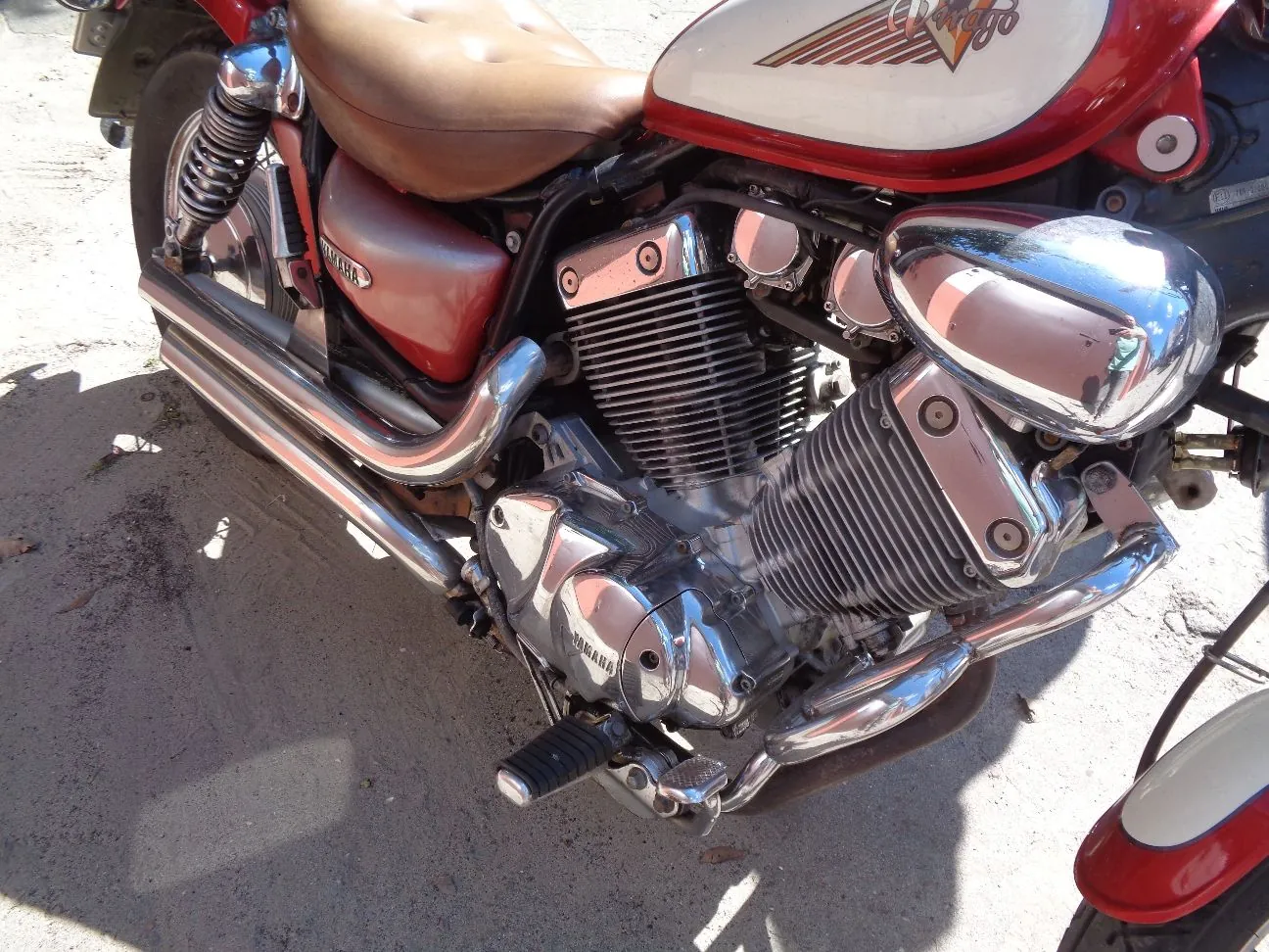 Zbliżenie na silnik i detale czerwonego motocykla marki Yamaha. Widoczny jest chromowany silnik z żebrowaniem, rury wydechowe, brązowe skórzane siedzenie oraz czerwony zbiornik paliwa z białym wstawką i napisem. Motocykl stoi na piaszczystym podłożu.