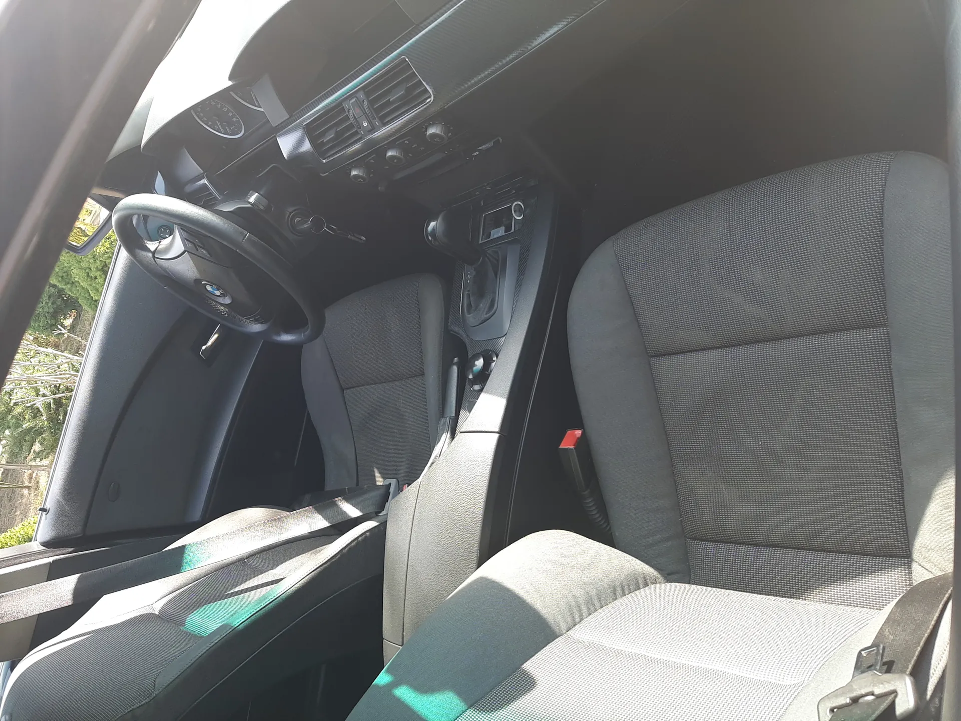 Wnętrze samochodu marki BMW z widoczną kierownicą z logo, szarymi siedzeniami tapicerowanymi tkaniną, deską rozdzielczą z przyciskami i wskaźnikami, dźwignią zmiany biegów oraz widokiem na zielone drzewa za oknem. Na siedzeniach widoczne są pasy bezpieczeństwa.