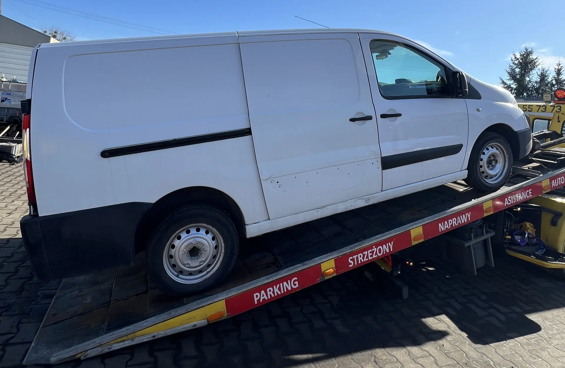 Biały furgon umieszczony na płycie holownika. Holownik posiada czerwono-żółte oznaczenia z widocznym tekstem: PARKING, STRZEŻONY, NAPRAWY, ASISTANCE, AUTO oraz numerem telefonu 55 73 73. Tło stanowi brukowana powierzchnia, budynek, drzewa i jasne niebo. Furgon ma czarne listwy dekoracyjne i srebrne felgi.