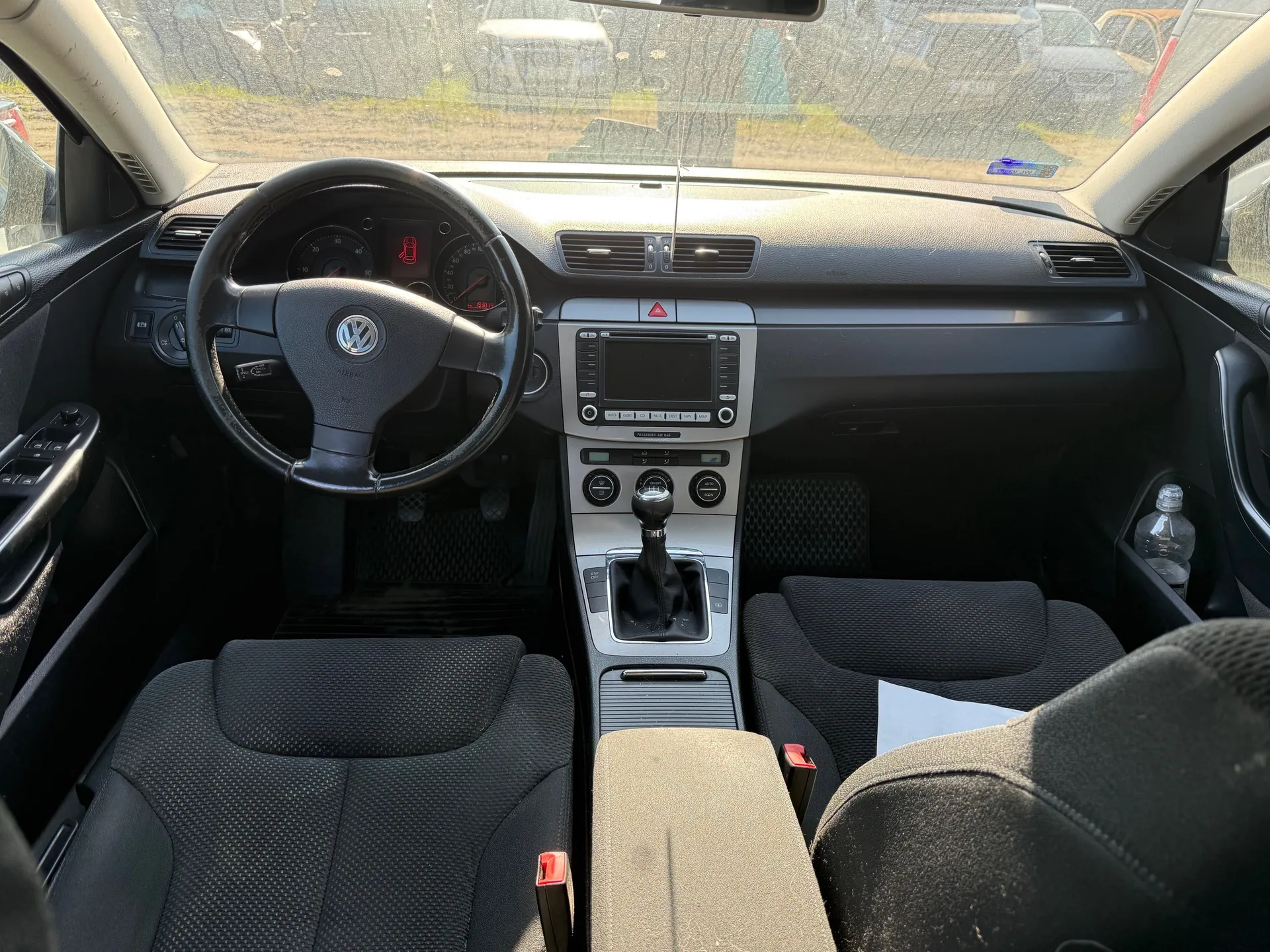 Wnętrze samochodu Volkswagen z widokiem na kierownicę z logo VW, czarną tapicerką siedzeń, centralną konsolą z ekranem, przełącznikiem biegów i licznikami. Na desce rozdzielczej widoczny czerwony sygnalizator. Przez przednią szybę widać inne pojazdy i teren z trawą. W otworze drzwi znajduje się butelka.