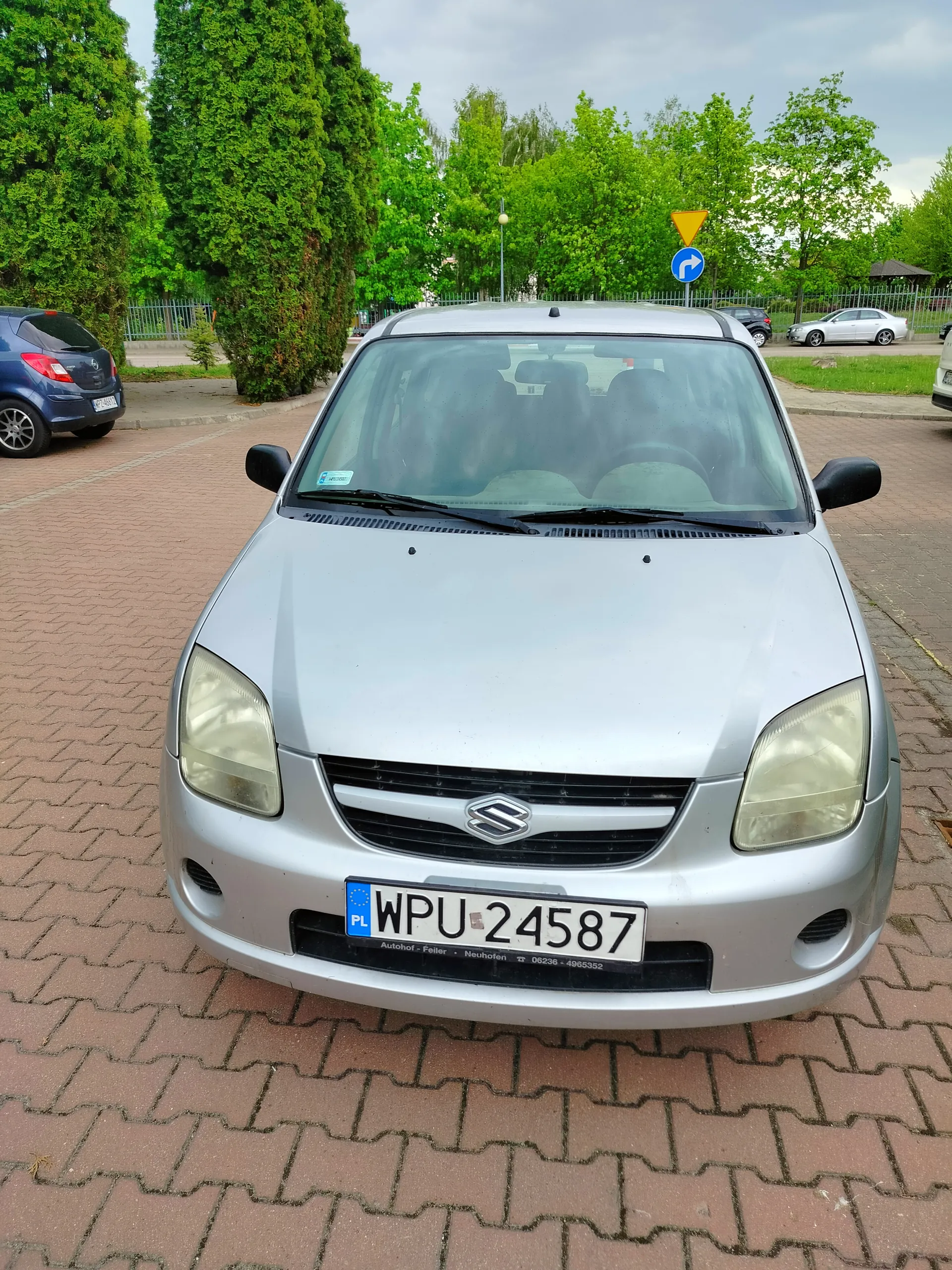Srebrny samochód Suzuki z tablicą rejestracyjną PL WPU 24587, zaparkowany na brukowanym placu. W tle widoczne są inne pojazdy, zielone drzewa, ogrodzenie oraz znaki drogowe: żółty trójkąt i niebieski okrąg z białą strzałką skręcającą w prawo. Na tablicy rejestracyjnej widoczny napis 'Autohof - Feller - Neuhofen 06236 - 4965352'.