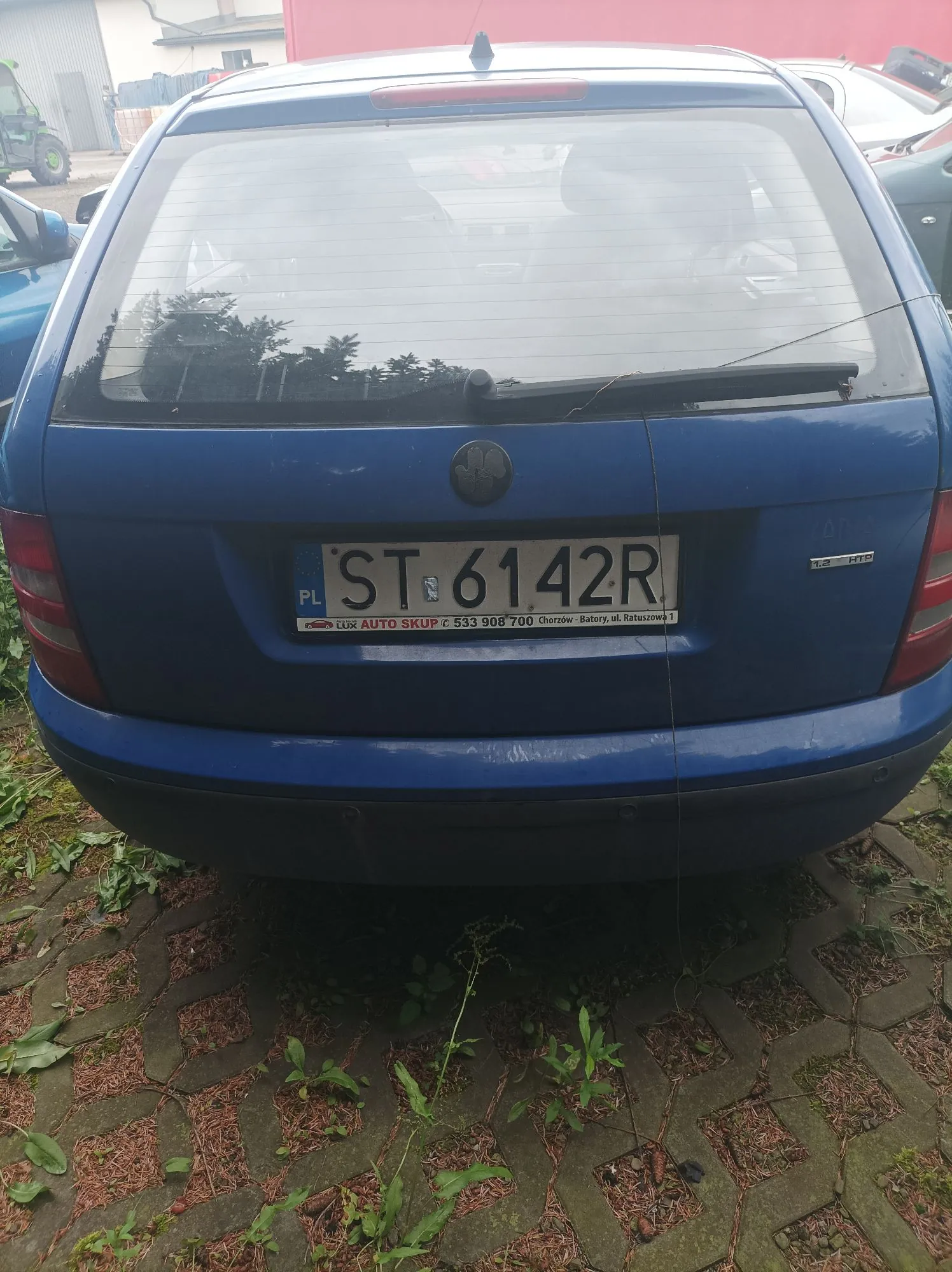 Tylny widok niebieskiego samochodu osobowego. Na klapie bagażnika znajduje się polska tablica rejestracyjna o numerze ST 6142R. Poniżej tablicy widnieje ramka z napisem: 'LUX AUTO SKUP ✆ 533 908 700 Chorzów - Baterie, ul. Ratuszowa 1'. Samochód stoi na kostce brukowej z wyrastającą między szczelinami trawą.