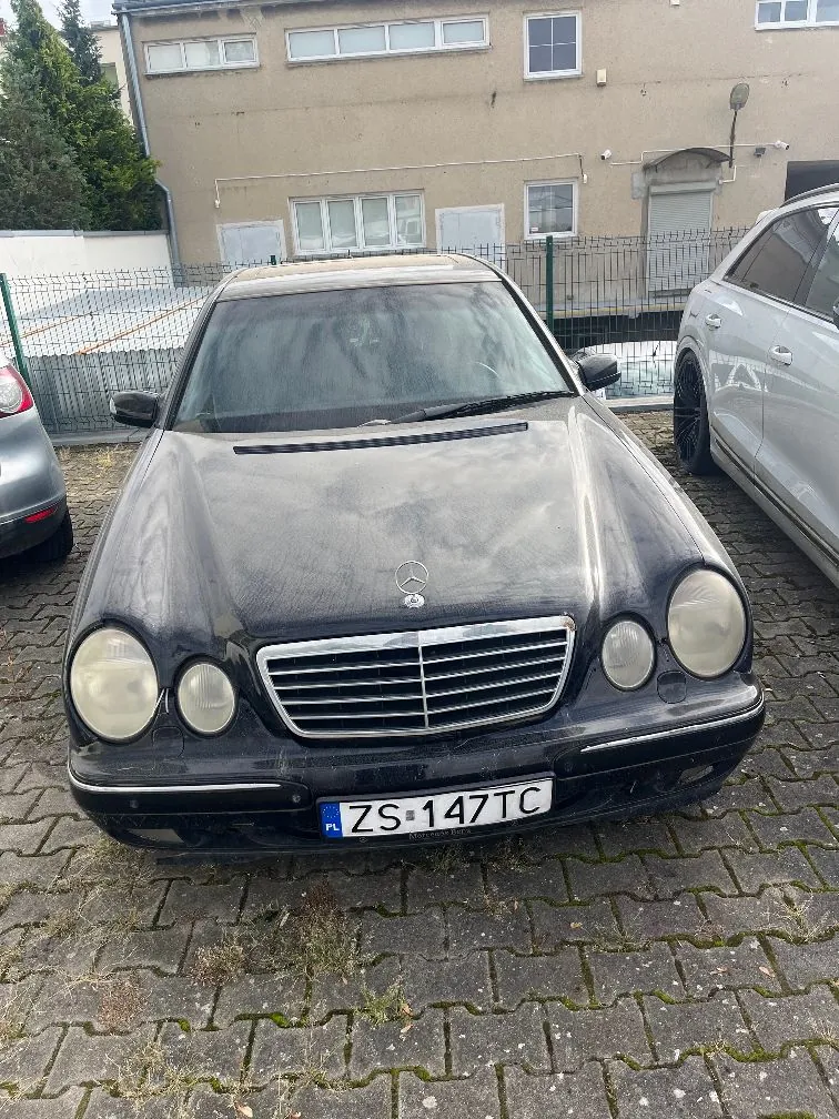 Czarny samochód marki Mercedes-Benz zaparkowany na kostce brukowej przed budynkiem. Auto jest zakurzone, posiada polską tablicę rejestracyjną o numerze ZS 147TC. W tle widoczny jest jasny budynek z oknami oraz fragment innego, srebrnego samochodu po prawej stronie.