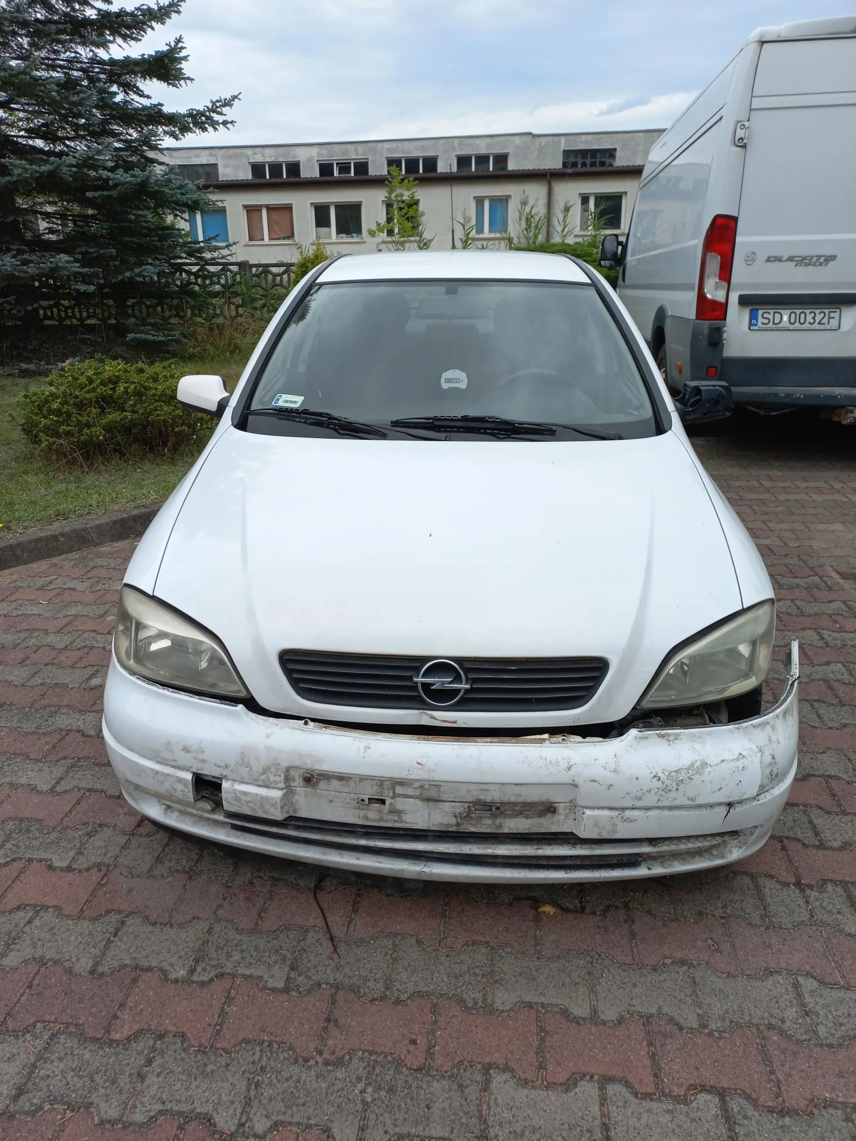 Biały samochód osobowy marki Opel zaparkowany na kostce brukowej. Przedni zderzak pojazdu jest uszkodzony, częściowo oderwany i brudny. W tle widoczny jest biały furgon marki Fiat Ducato z tablicą rejestracyjną SD 0032F oraz jasny budynek mieszkalny i drzewa.