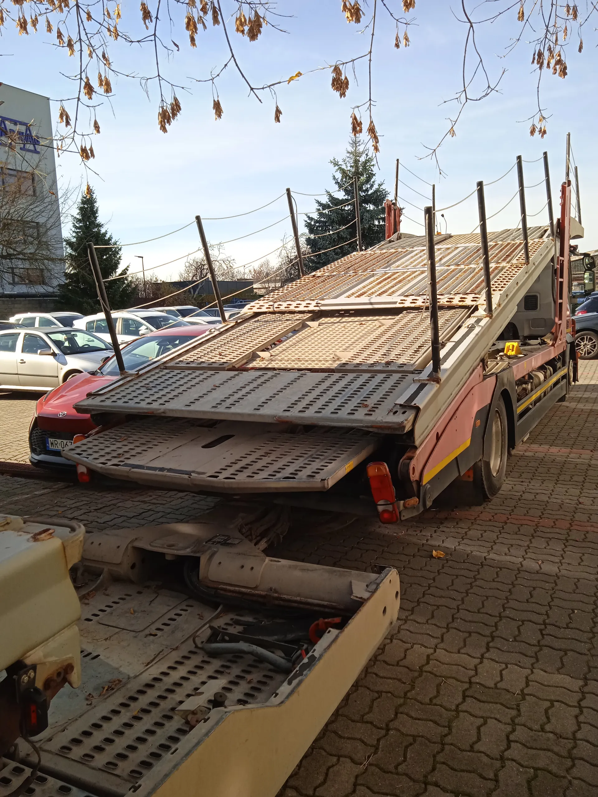 Samochód transportowy do przewozu pojazdów z rozłożonym rampą, zaparkowany na brukowanej powierzchni. W tle czerwony samochód Toyota z tablicą rejestracyjną WR 047, inne zaparkowane pojazdy, budynek z niebieskim napisem oraz drzewa bez liści. Jasne niebo z widocznymi gałęziami na pierwszym planie.