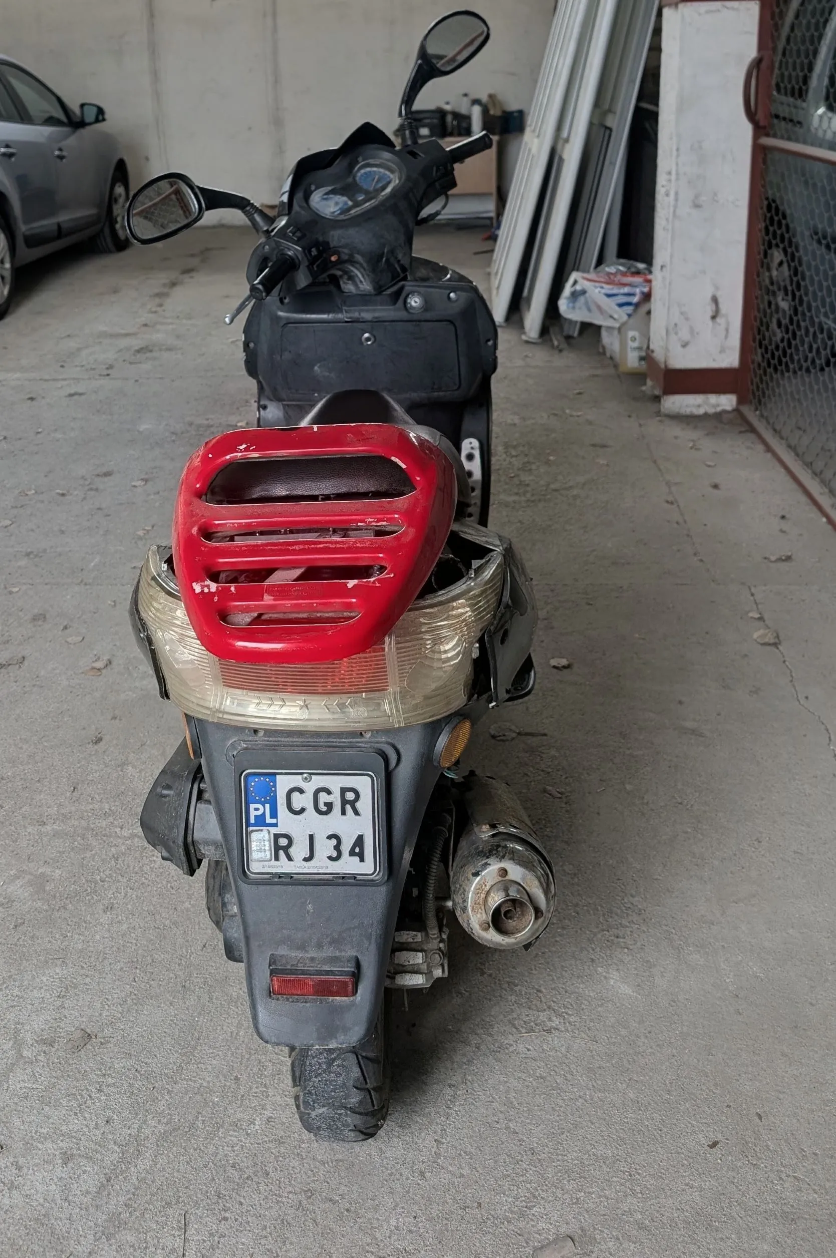 Widok z tyłu na czarny skuter zaparkowany w garażu lub warsztacie. Pojazd ma czerwoną obudowę tylnej lampy, która jest częściowo uszkodzona. Na skuterze znajduje się polska tablica rejestracyjna o numerze CGR RJ34. W tle widoczny jest fragment srebrnego samochodu oraz oparte o ścianę białe panele.