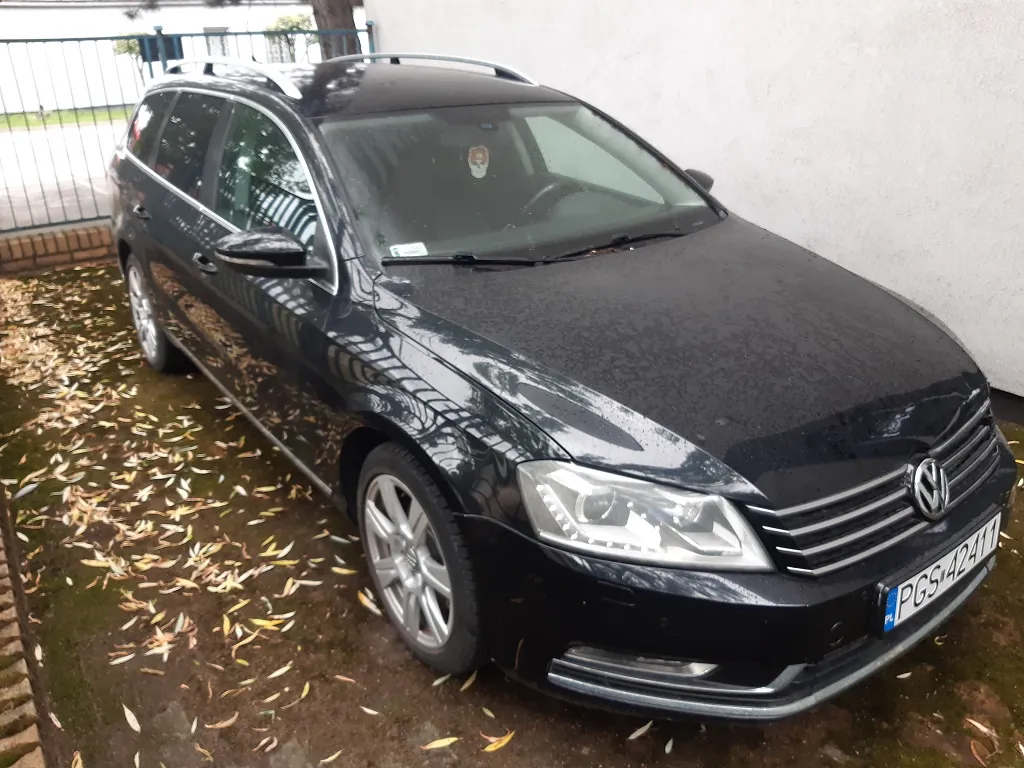 Czarny samochód marki Volkswagen w nadwoziu kombi, zaparkowany na podjeździe wysypanym liśćmi obok białej ściany budynku. Auto posiada srebrne relingi dachowe i felgi aluminiowe. Tablica rejestracyjna ma numer PGS 4241T.