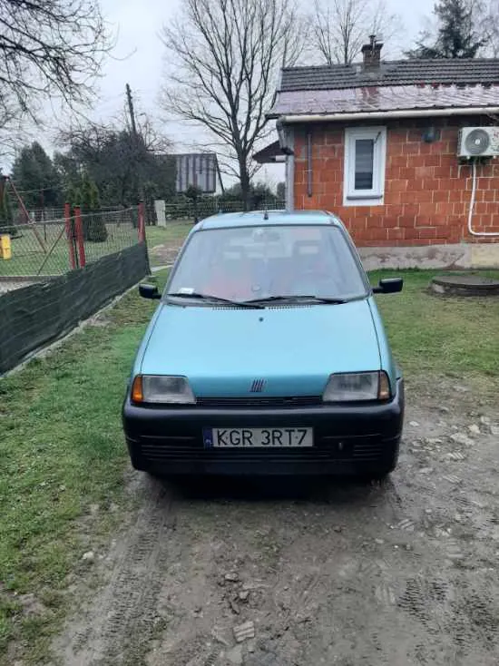 Niebieski samochód osobowy zaparkowany na ziemnej drodze przed ceglanym domem z białym oknem i klimatyzatorem. Na tablicy rejestracyjnej widoczny napis KGR 3RT7. W tle drzewa bez liści, ogrodzenie z czerwonymi słupkami oraz zielona trawa. Niebo jest zachmurzone.